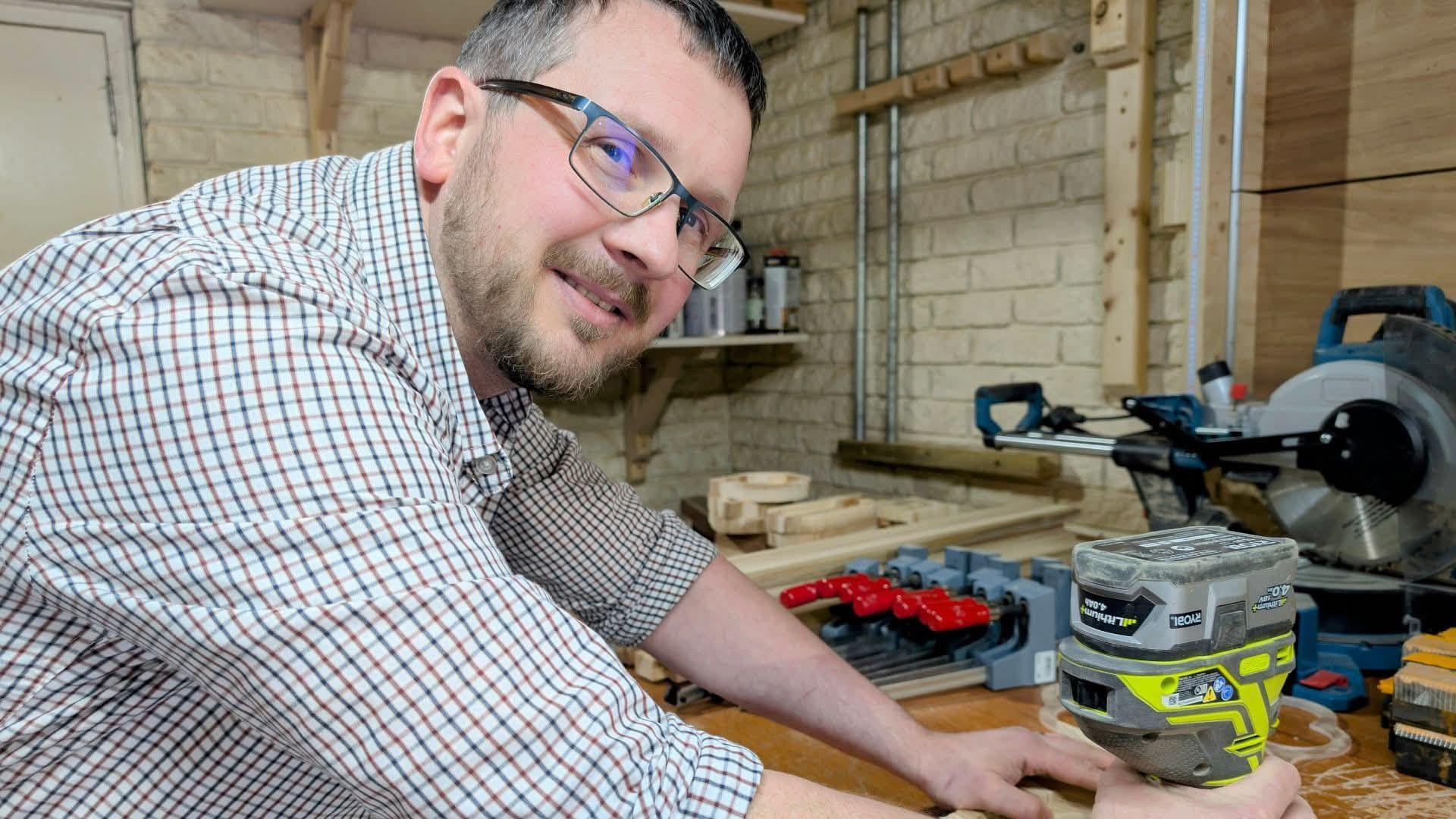 Bob working on a piece of wood, sanding it down in his workshop. He is looking at the camera and has short grey hair and beard, wearing thin framed glasses and a checkered shirt. 