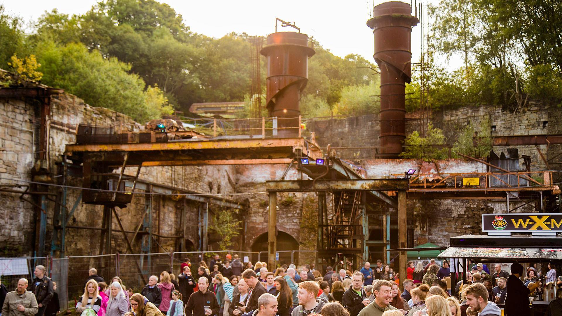 Crowds gathered on the Brymbo site in Wrexham. The framework of a steelworks can be seen above the crowd, along with green forestry behind the walls of the unit. 