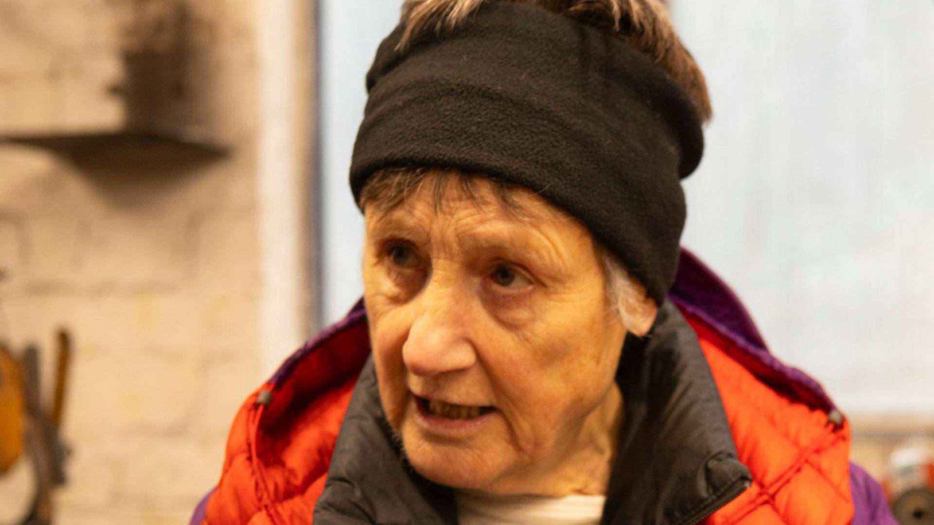 Roz Wollen, a woman wearing a black headscarf and a red coat. She is in an industrial building looking away from the camera.