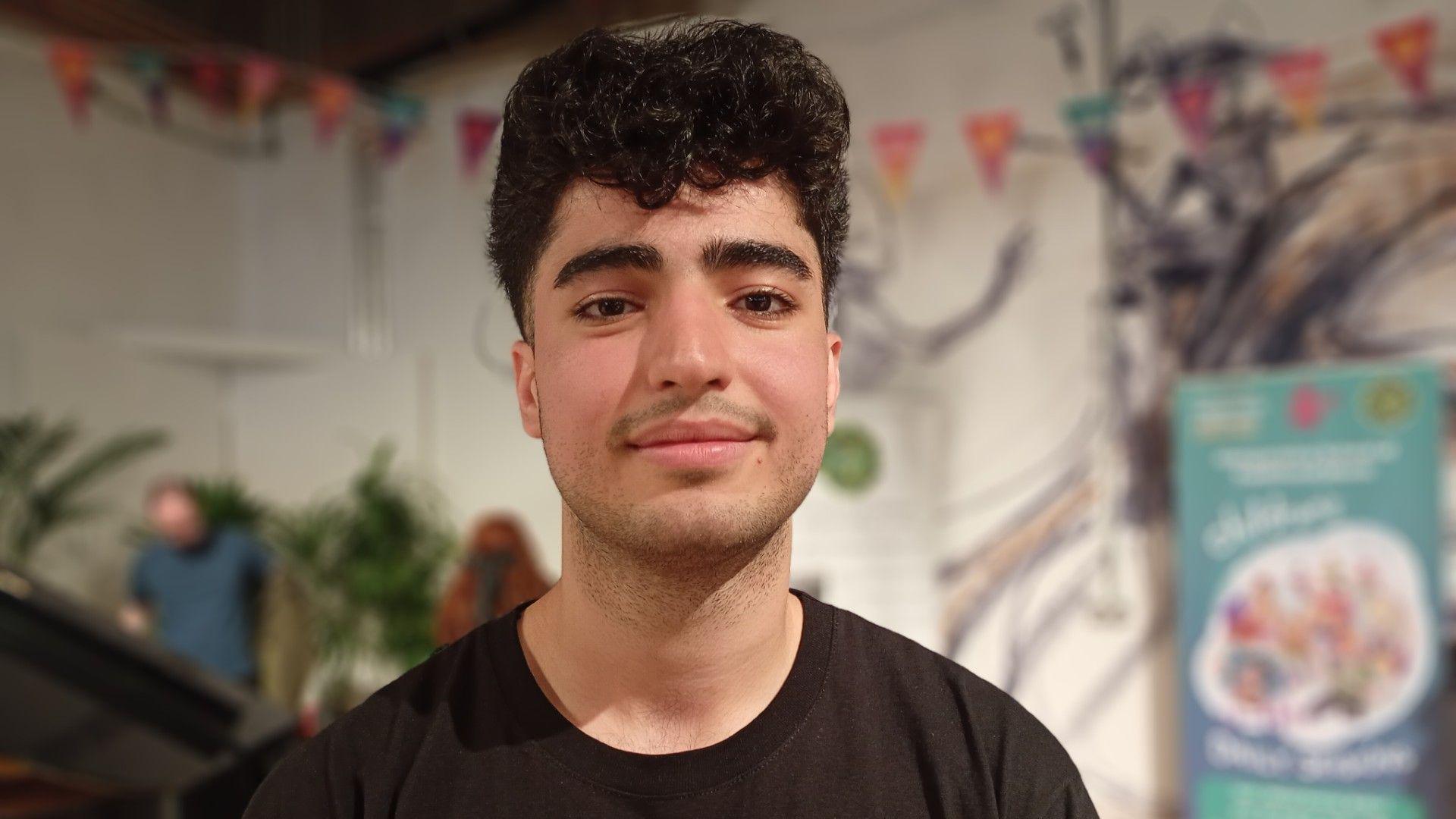 A man with dark hair looking at the camera, he is wearing a black top