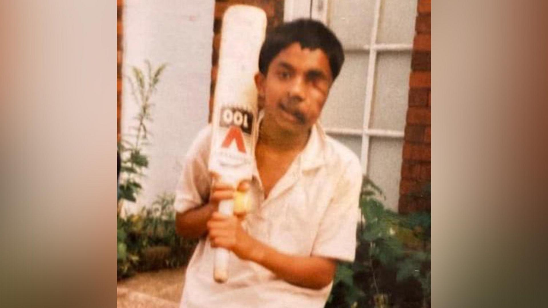 A boy with dark hair and wearing cricket whites holds a cricket bat over his shoulder.