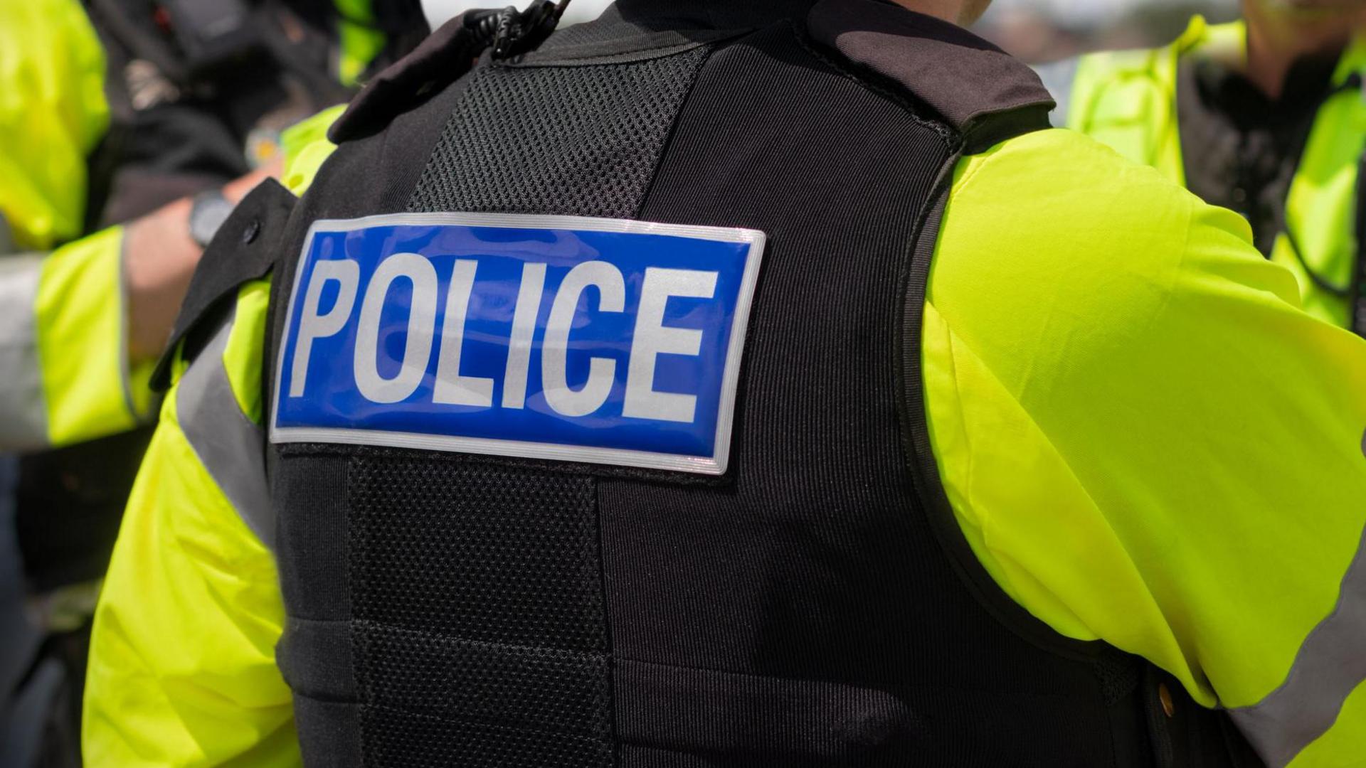 A close-up of 'POLICE' marking written on the back of a hi-visibility stab proof vest worn by a trio of police officers at the scene of an incident.