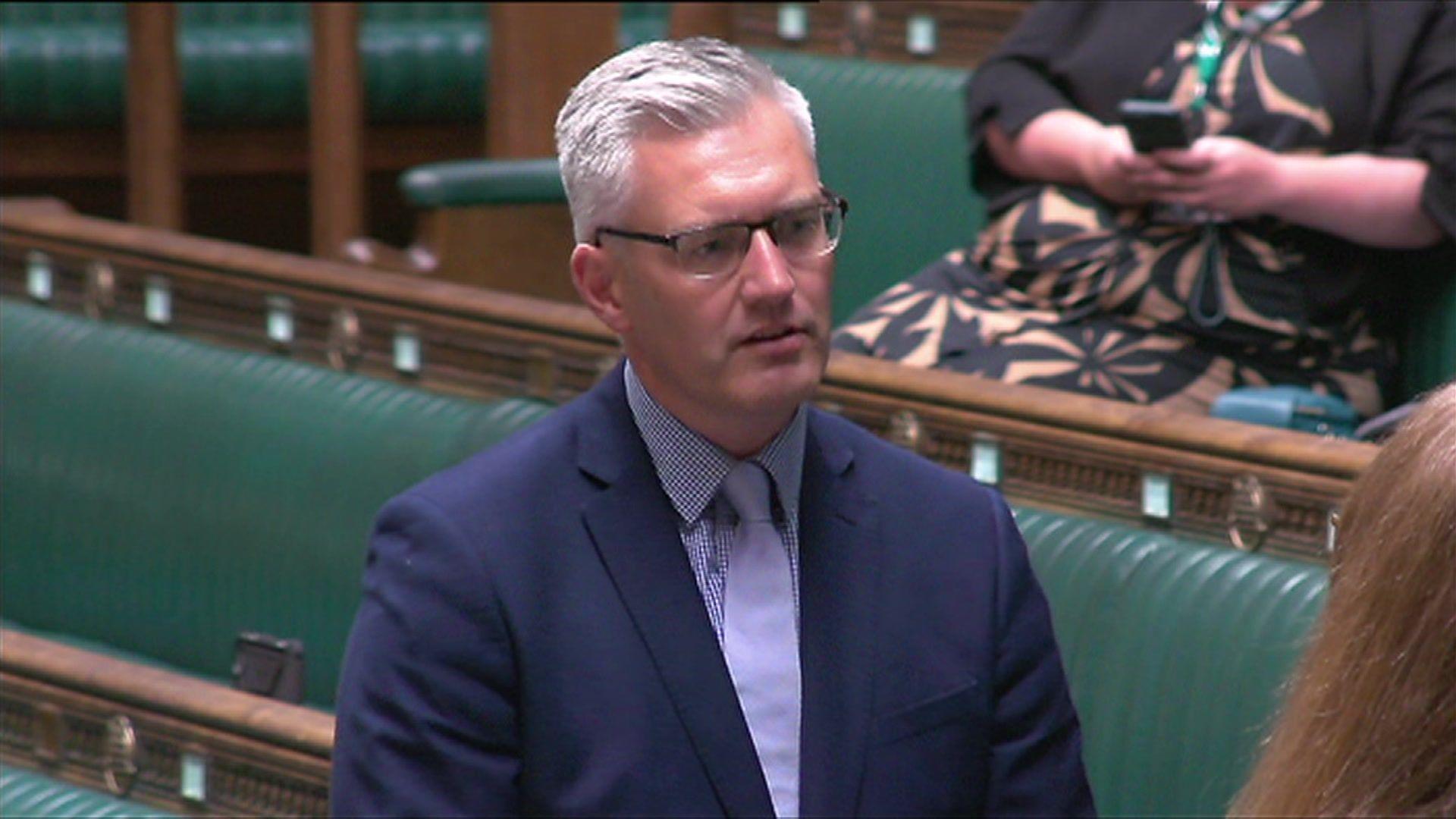 David Smith in the House of Commons. He has short grey hair and is wearing glasses. He has a navy blue suit, with a blue shirt and tie. Behind him are the leather benches where MPs sit.