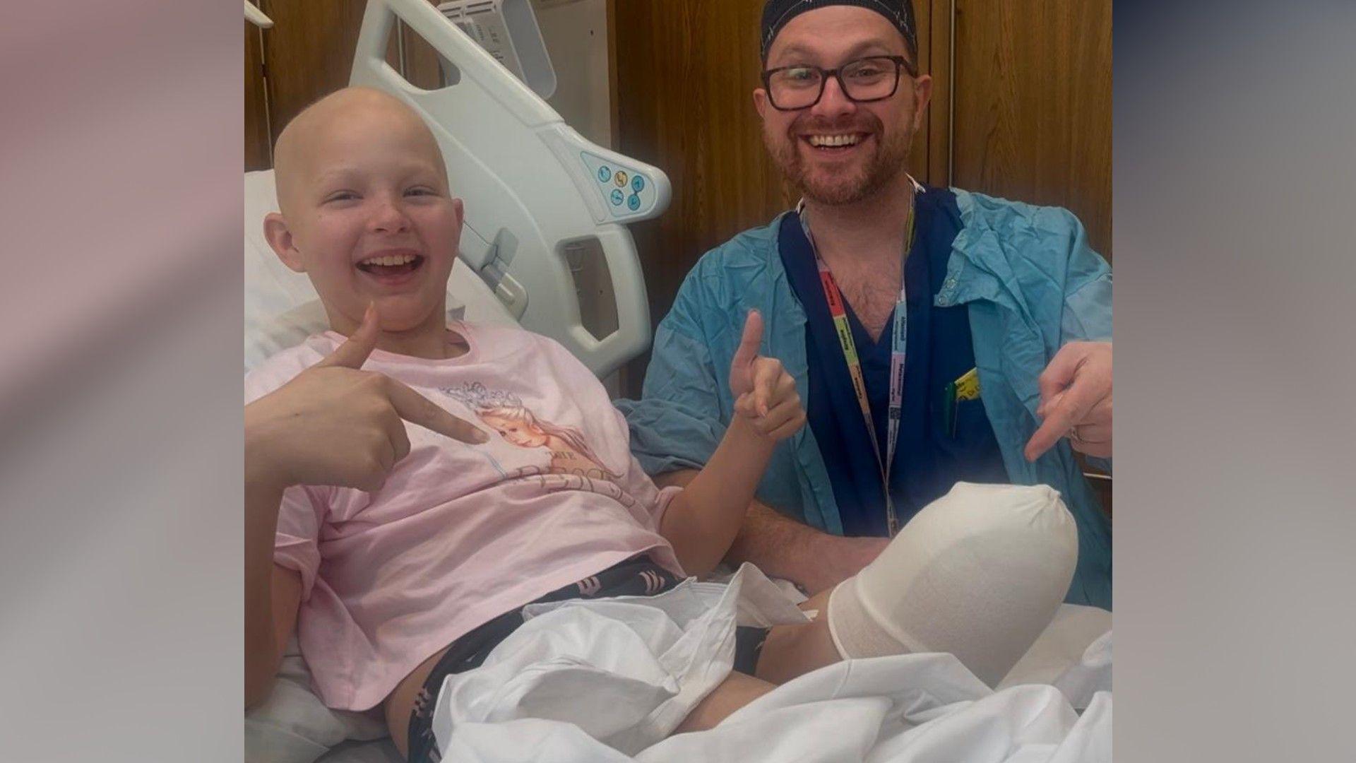 Millie wearing a pink T-shirt - sitting in a hospital bed - smiling and pointing to her bandaged and amputated leg, with a doctor smiling next to her hospital bed