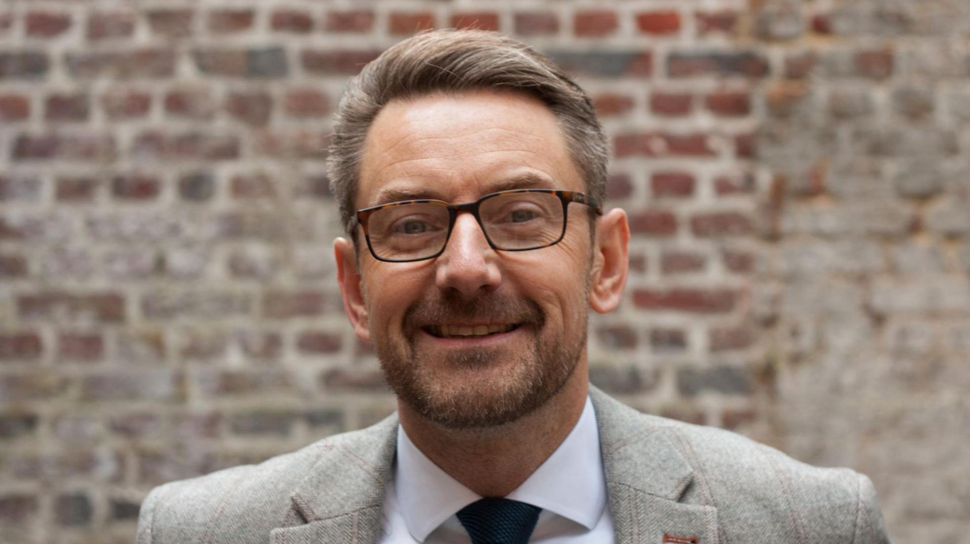 Malcolm Fleming is smiling at the camera. He has a stubble beard and short grey/brown hair. He is wearing a grey blazer and a shirt and tie.
