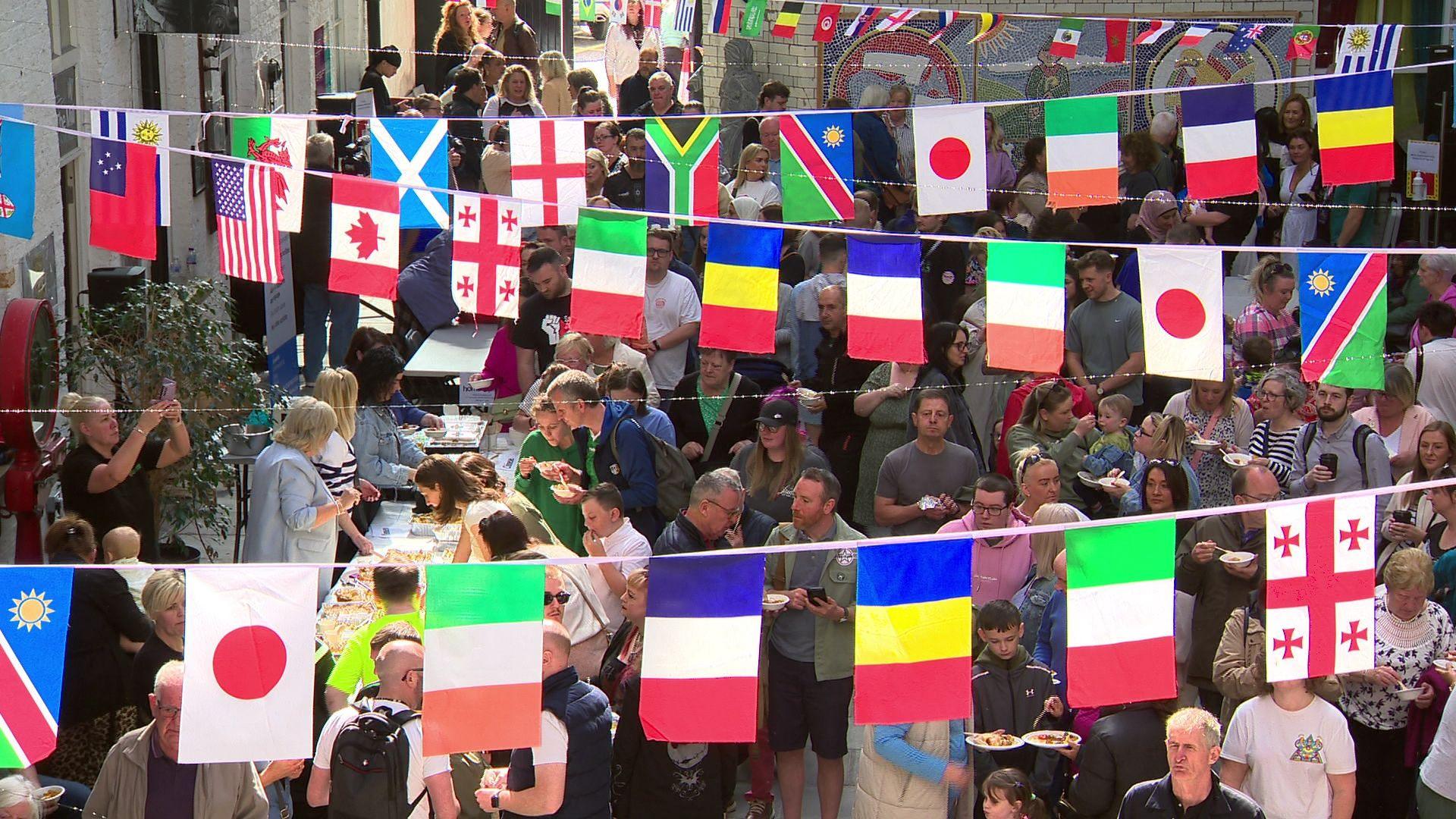 A large crowd of people queue by food stalls at the Féile an Phobail in west Belfast. Flags from around the world hang above them.