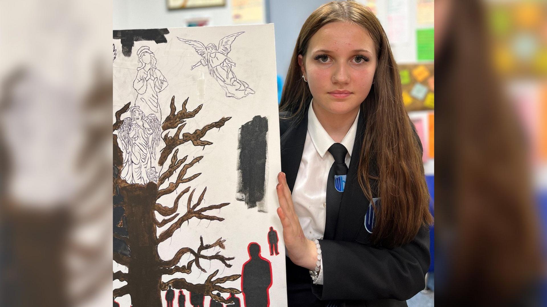 A teenage girl with long brown hair wearing a black school uniform holds up a canvas with a painting of what appears to be an angel on top of a tree.