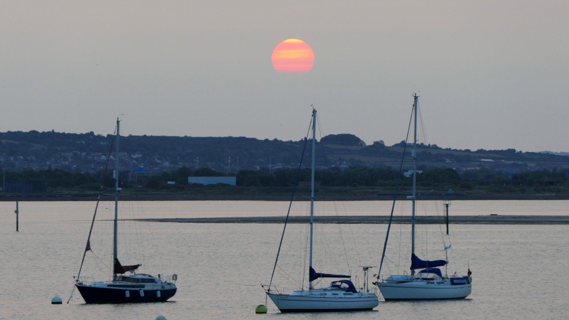 Orange disc of the sun set against brown tinged skies. Water and boats in the foreground.