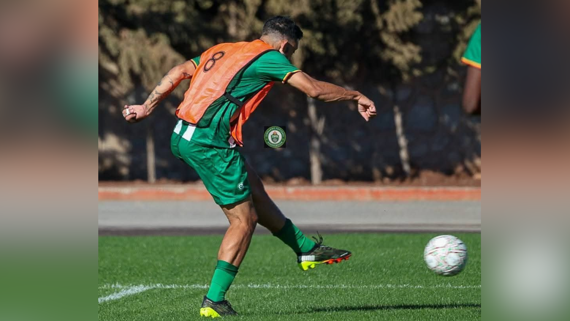 Bonne seen from the side kicking a white football with his left foot. He wears a green kit (T-Shirt and shorts) with an orange vest over the top, which has the number 8 in black. He wears green socks and has a tattoo on his left arm.