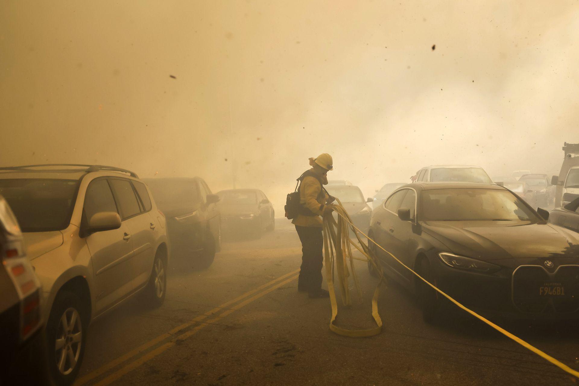 A Los Angeles firefighter drags a hose past cars trying in a smoky street in Pacific Palisades.