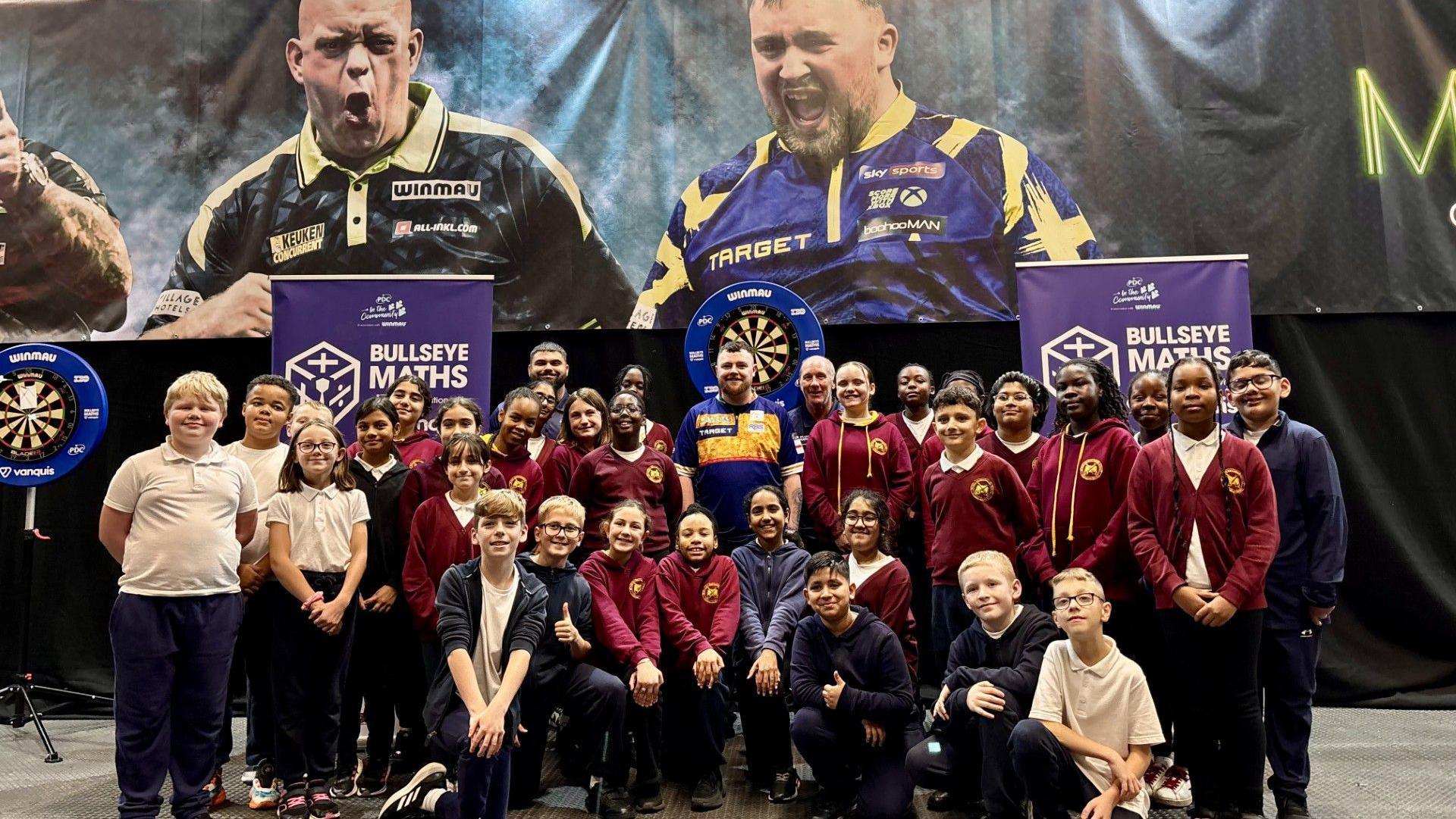 A group of school children standing infront of darts boards and some adults are also in the photo. They are smiling and some have their thumbs up. Their school uniform is maroon with a yellow crest.