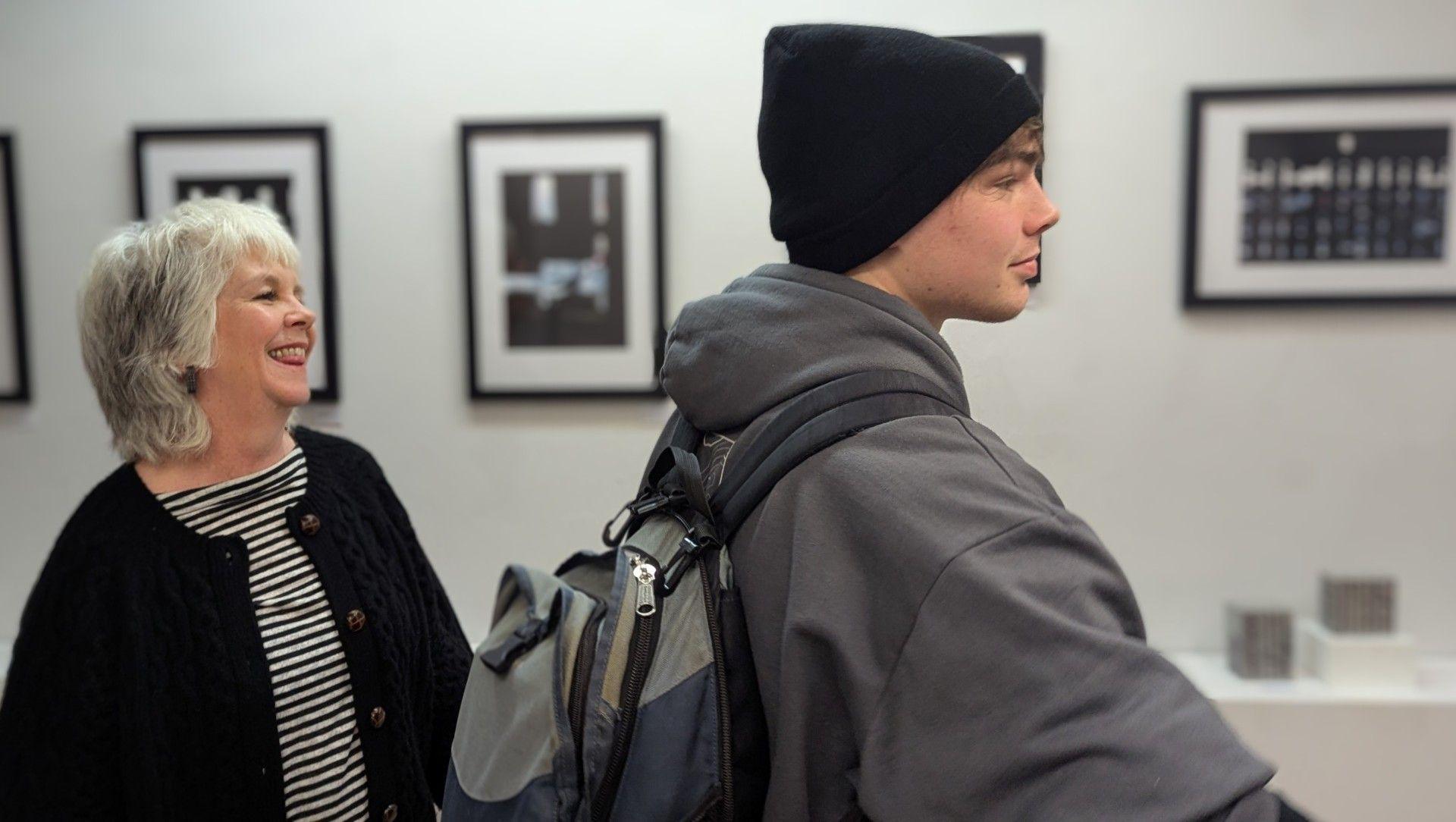 Alison Waters and her nephew Dylan look in sideprophile looking at the pictures and sculptures. Ms Waters has medium length grey hair, she wears a striped top and a black cardigan. Dylan wears a black beanie hat, a grey hoodie and a grey and blue rucksack.