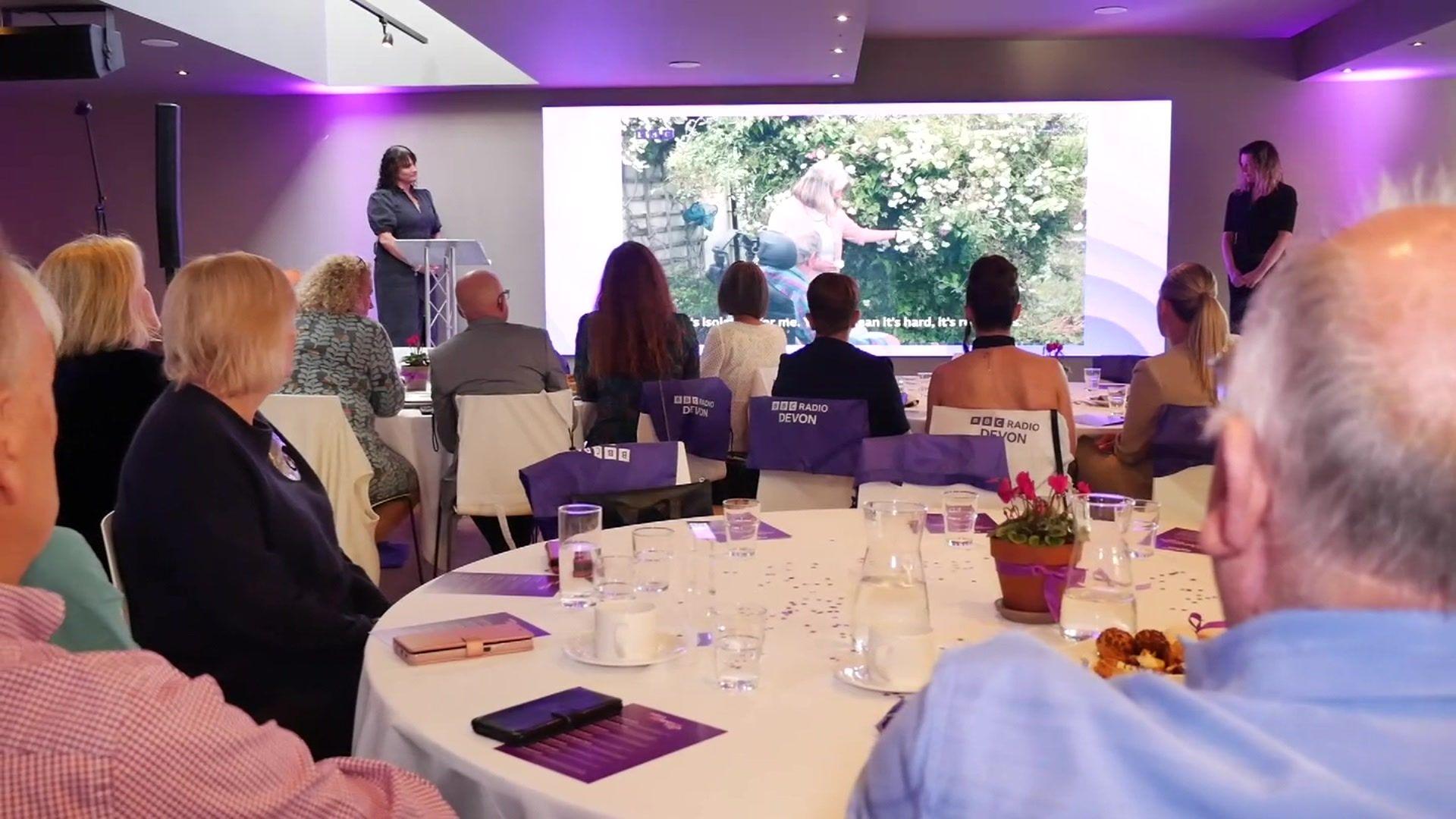 People are sat around round tables which have glasses of water and cups of tea on them. They are looking forward to a stage which has a screen which is playing a video. 