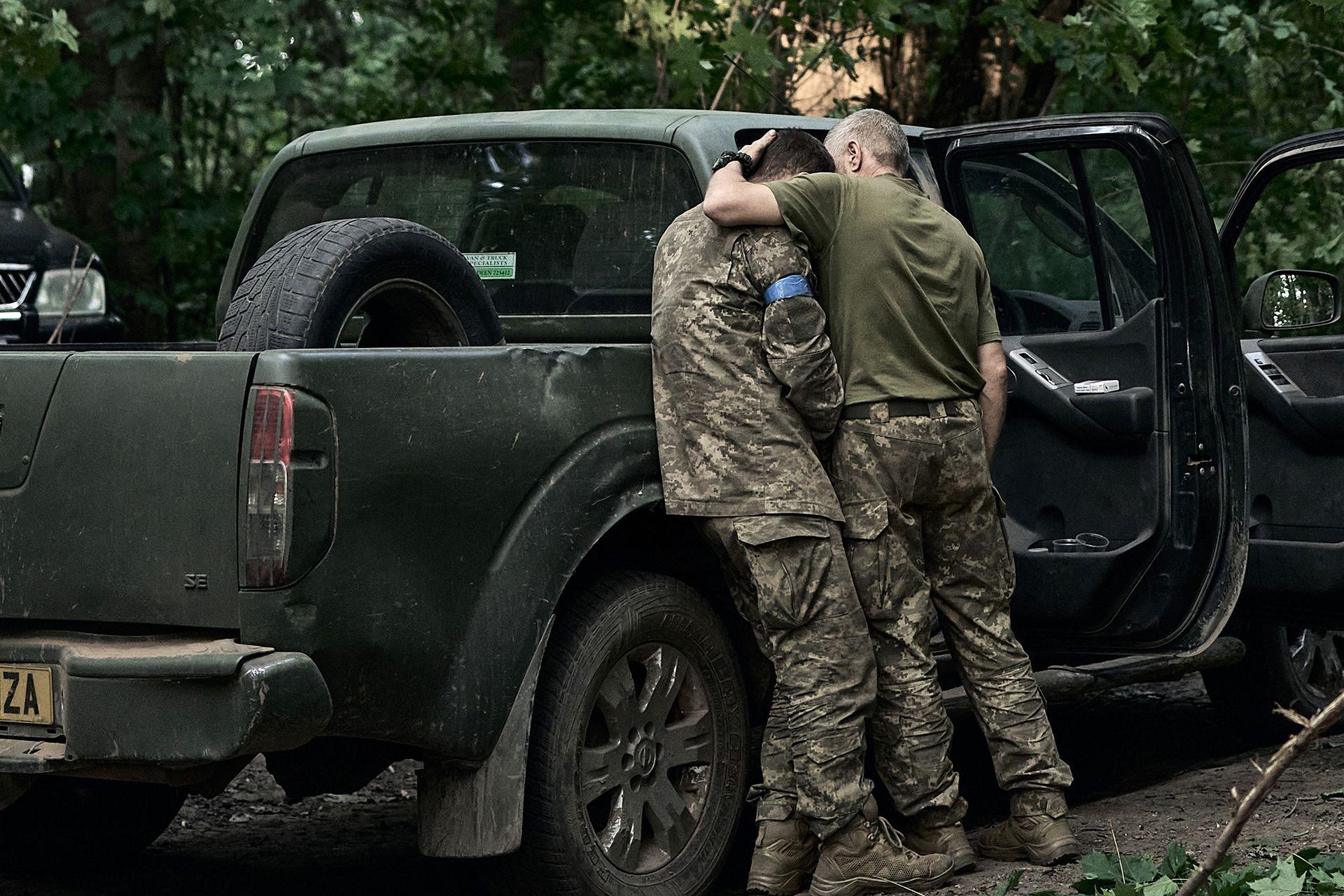 Two soldiers embrace, with one resting his head on the other's shoulder, both dressed in military outfits and standing next to a truck with its doors open and with bullet marks on its side, in the north-eastern Sumy region of Ukraine on 14 August 2024.