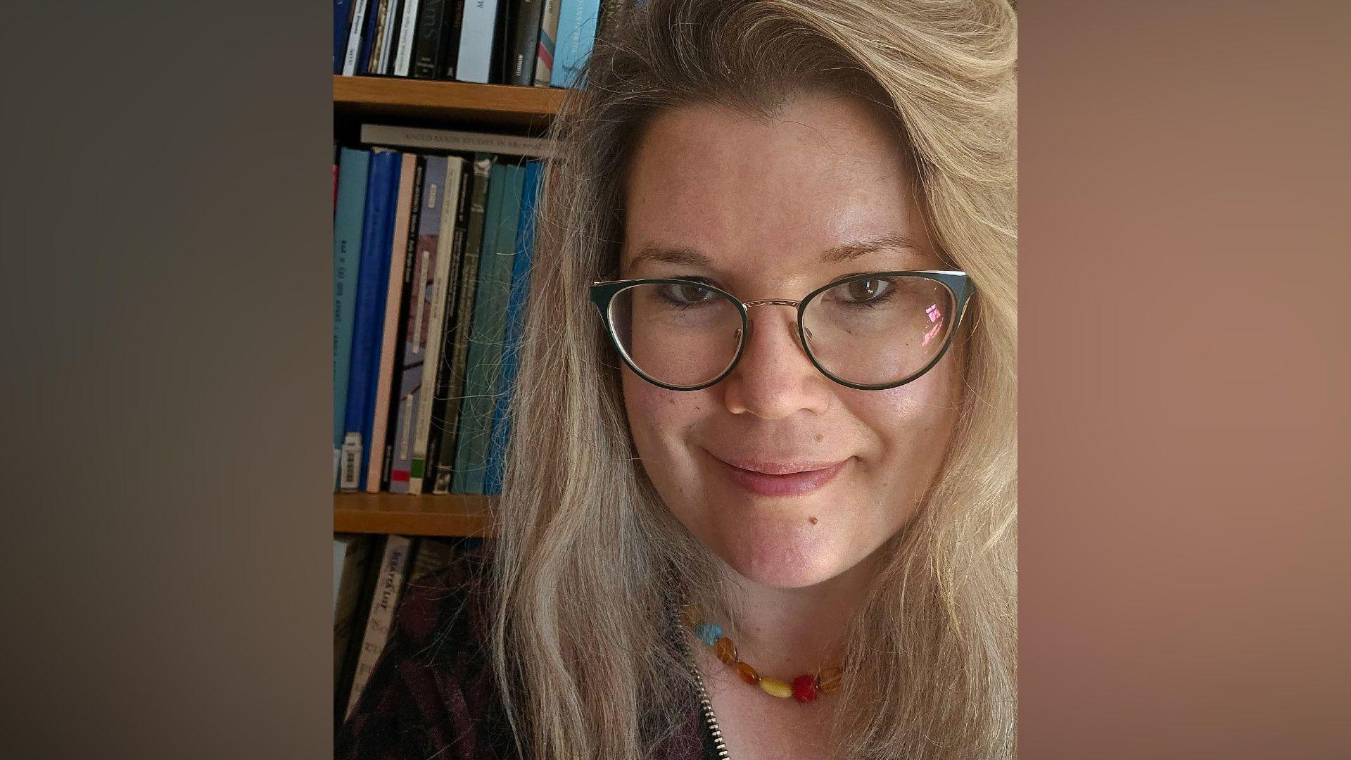 Një grua me syze dhe flokë të gjatë bjondë duke pozuar për një foto. Ajo mban një gjerdan shumëngjyrësh dhe qëndron përpara një rafti librash.