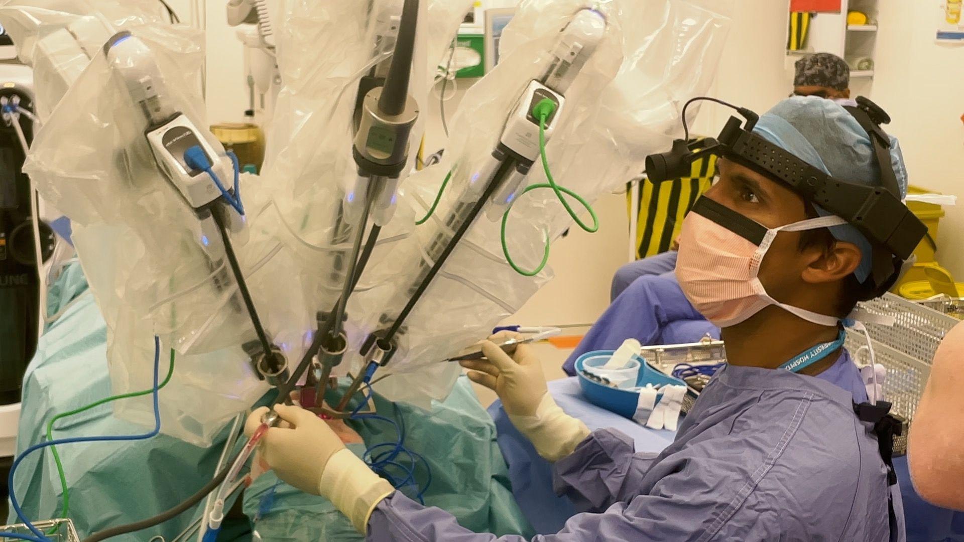 A surgeon in an operating theatre  wearing scrubs and surgical head wear. They are monitoring three robotic arms that are operating on a patients throat cancer.  