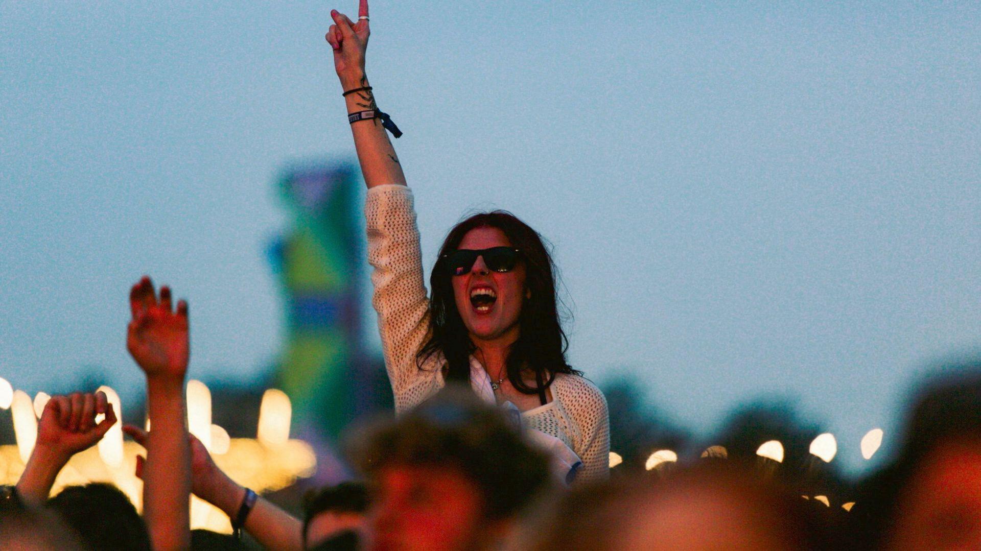 A festival goer is on someone's shoulders watching a performance. They have an arm reaching up into the air and have black shoulder length hair, wear black square framed glasses and a white crochet-knit long sleeved top. The background is blurred but shows the warmth of fairy lights.