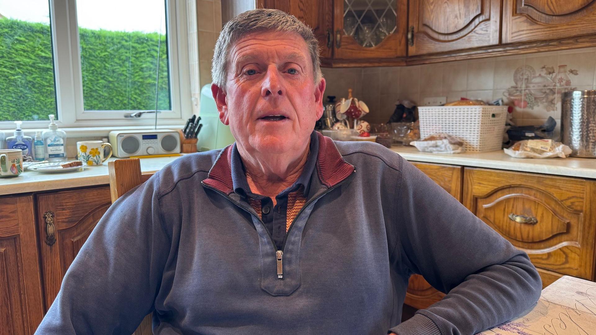 Gerry White sits at his kitchen table, he has short grey hair and is wearing a blue quarter-ziip top with maroon detailing on the inside of the collar. Behind him are brown wood kitchen cabinets and items including a radio, block of knives and bread basket on top of the kitchen counters.