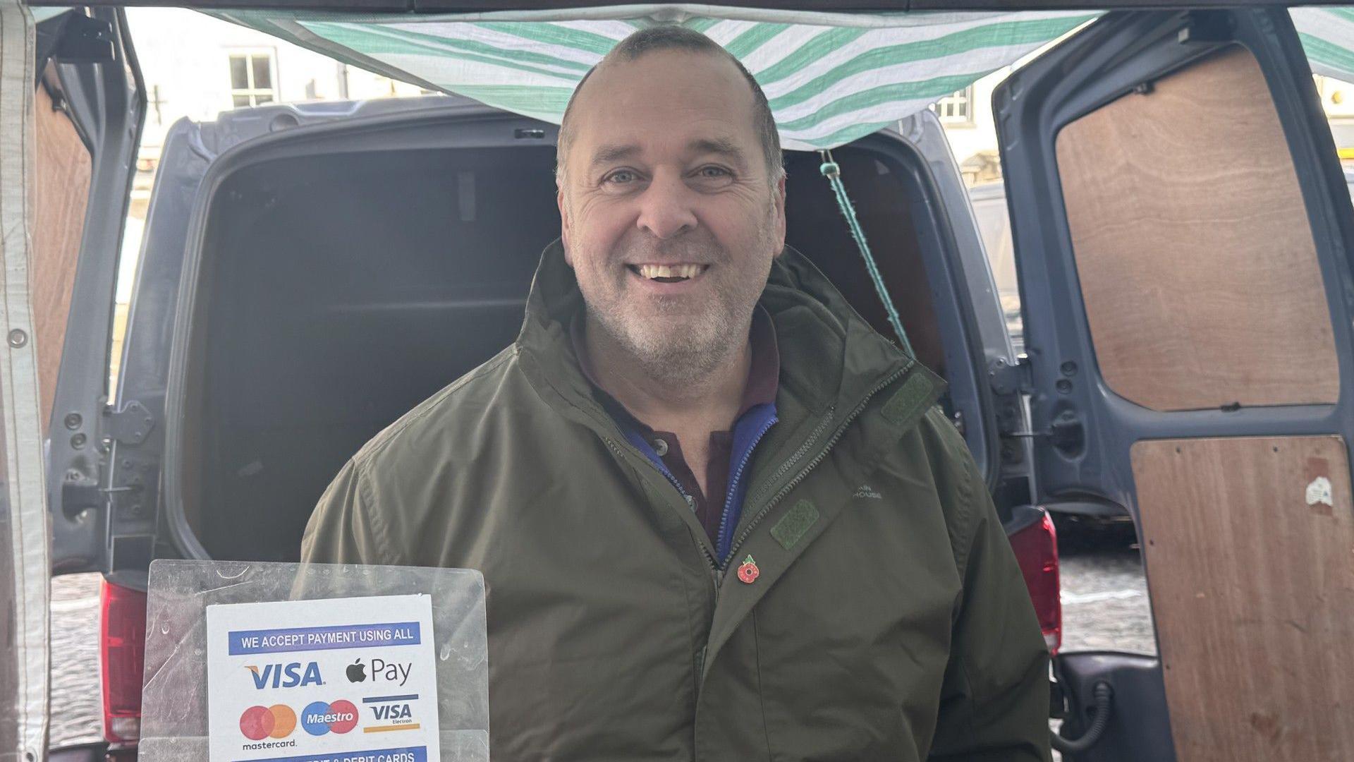 A middle aged man is smiling at the camera. He's wearing a green coat, with a blue zip up jumper underneath. He has a red poppy pin on, and is standing in front of the boot of a gray van, the doors open. A green and white stripe canopy is above him, and there's a sign detailing the payment types accepted to the left of the image.