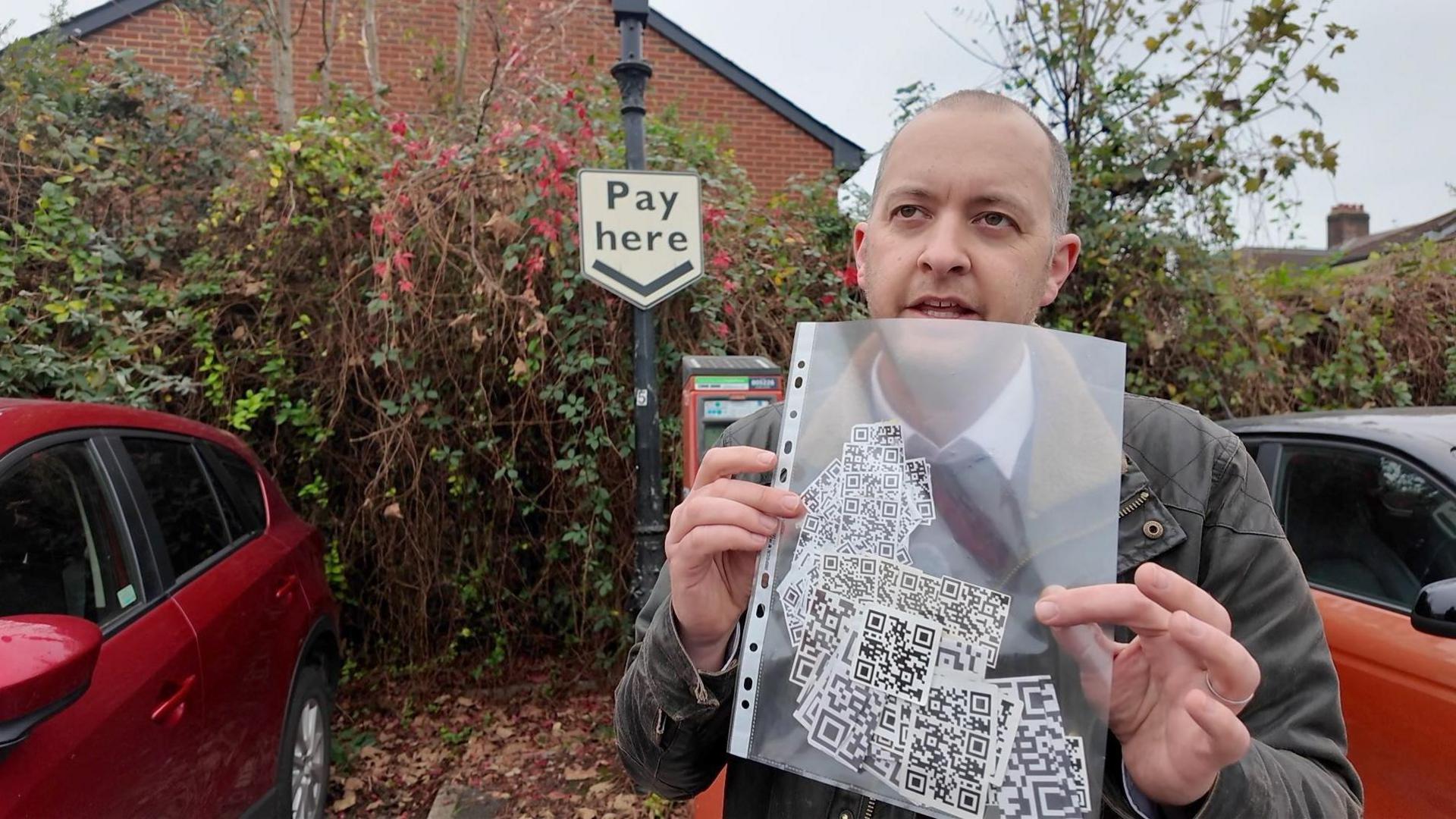 Richard Horton holding a see-through A4 sleeve full of QR code stickers in a car park. Behind him are two cars and a ticket machine with a 'Pay here' sign above it. 