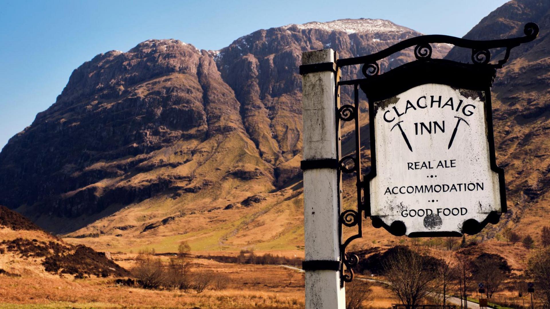 A sign for Clachaig Inn with towering mountains in the background. The metal sign reads: "Clachaig Inn, Real ale. Accommodation. Good food." The word "inn" has an ice axe either side of it.