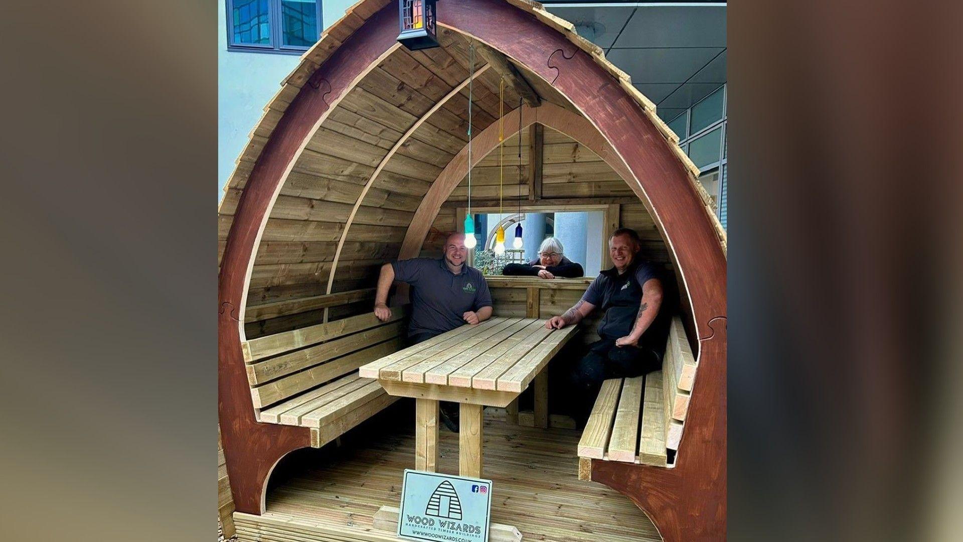 A triangular wooden seating pod with a bench and two men sitting on either side of the bences. Both are wearing black and smiling at the camera.
There is a third person also in the photo peering through the window of the pod.