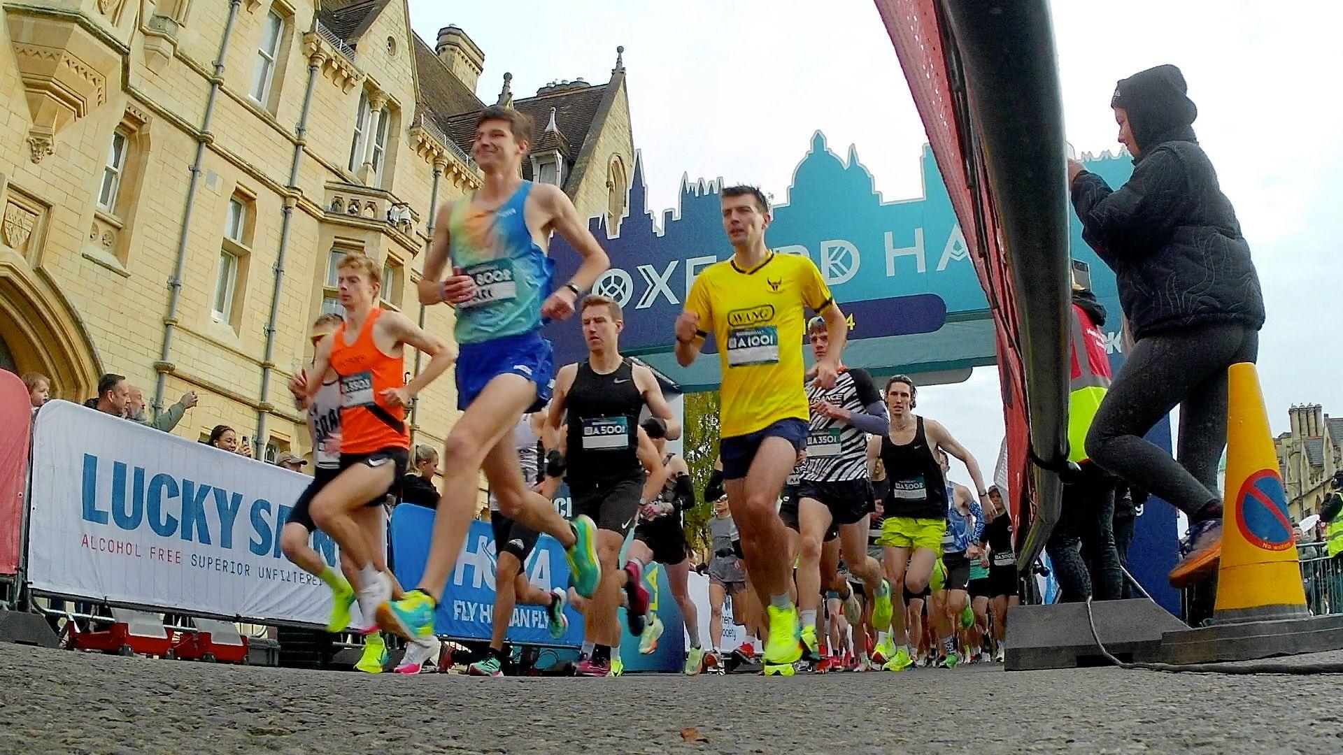 A large pack of runners with advertising boards to either side of them. behind them is a big blue archway saying 'Oxford Half' in white letters.