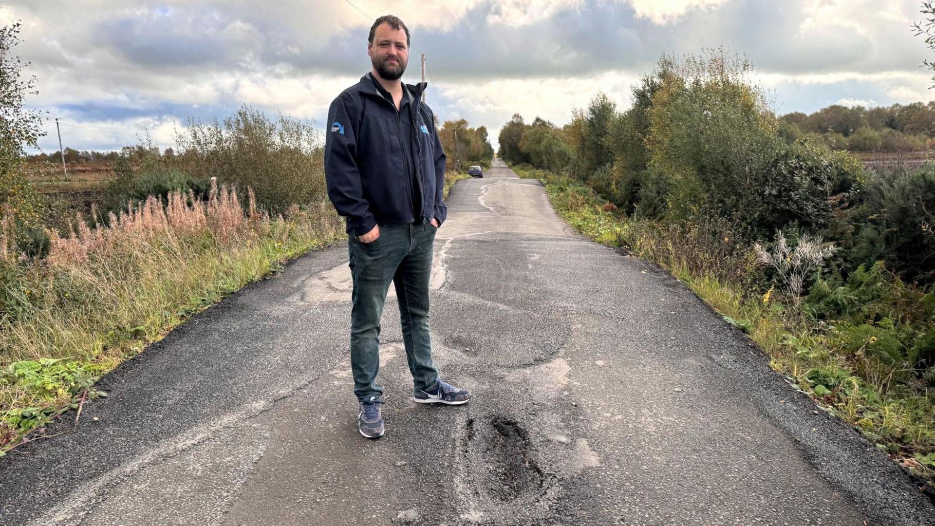 Malachy Quinn stands in the middle of a small rural road during the daytime. The road is lined with greenery and trees. He is standing with his hands in his pockets in front of a small pothole