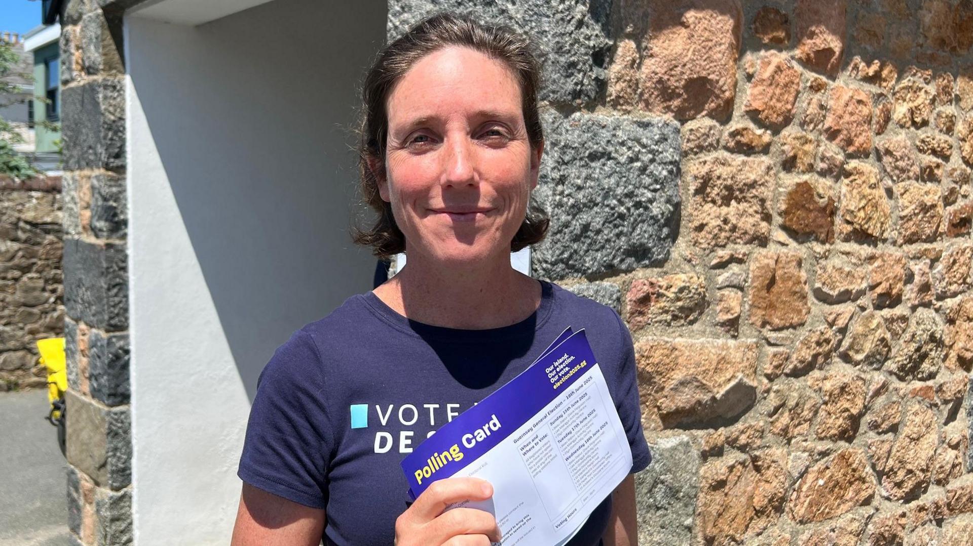 A short-haired woman in a blue T-shirt, holding polling card in the sun while she smiles.