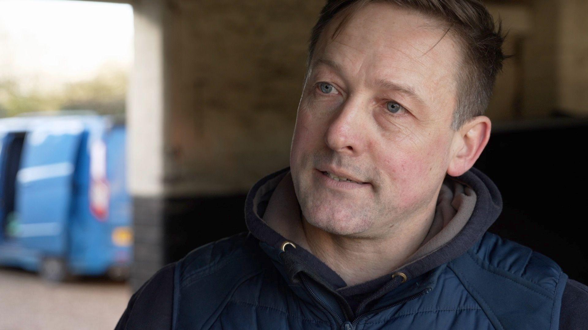 Middle-aged man with short brown hair wearing a blue gilet with a blue van in the background.