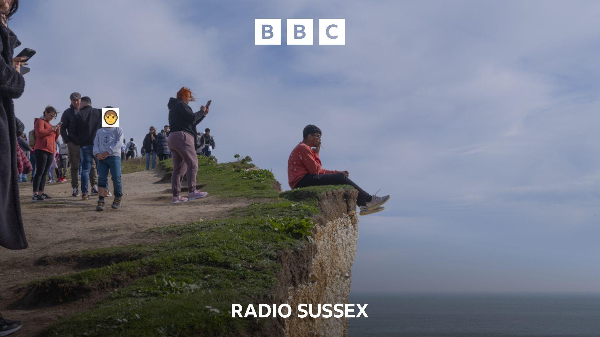 A large number of visitors stand near a chalk cliff edge overlooking the sea. One man in a red top sits with his legs dangling over the edge.