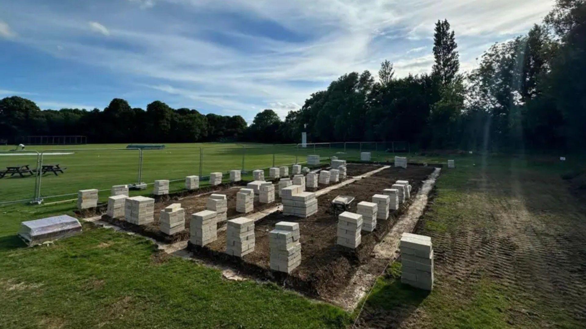 The cricket field with stacks of raw materials around a fenced area- in preparation of the new pavilion works 