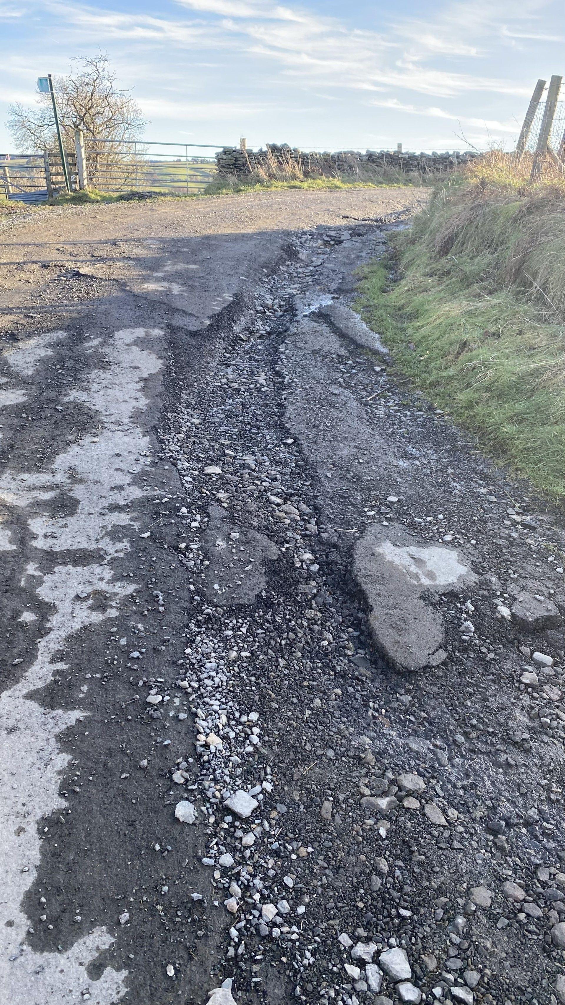 A picture of a  country lane which has a long and narrow pothole and loose stones, a field gate and a footpath sign are visible in the background