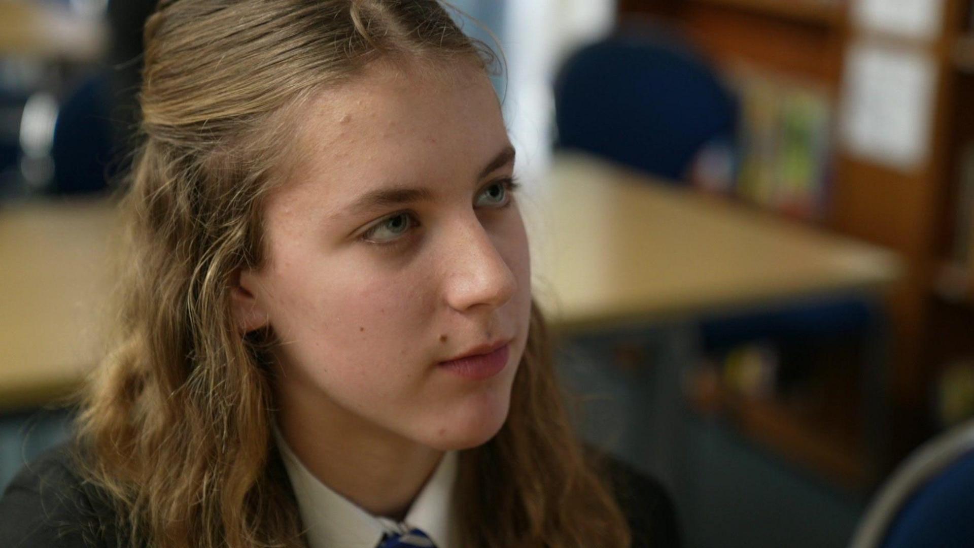 Sophia has long blonde hair and is wearing a school blazer and white shirt as she sits in front of a row of desks.