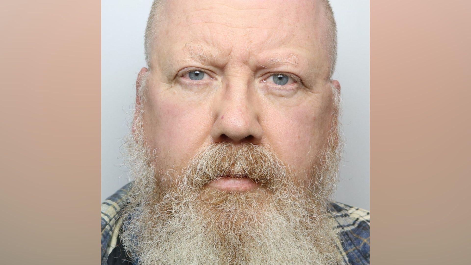 A man with a bald head and long grey beard. He is wearing a blue checked shirt and standing against a grey background
