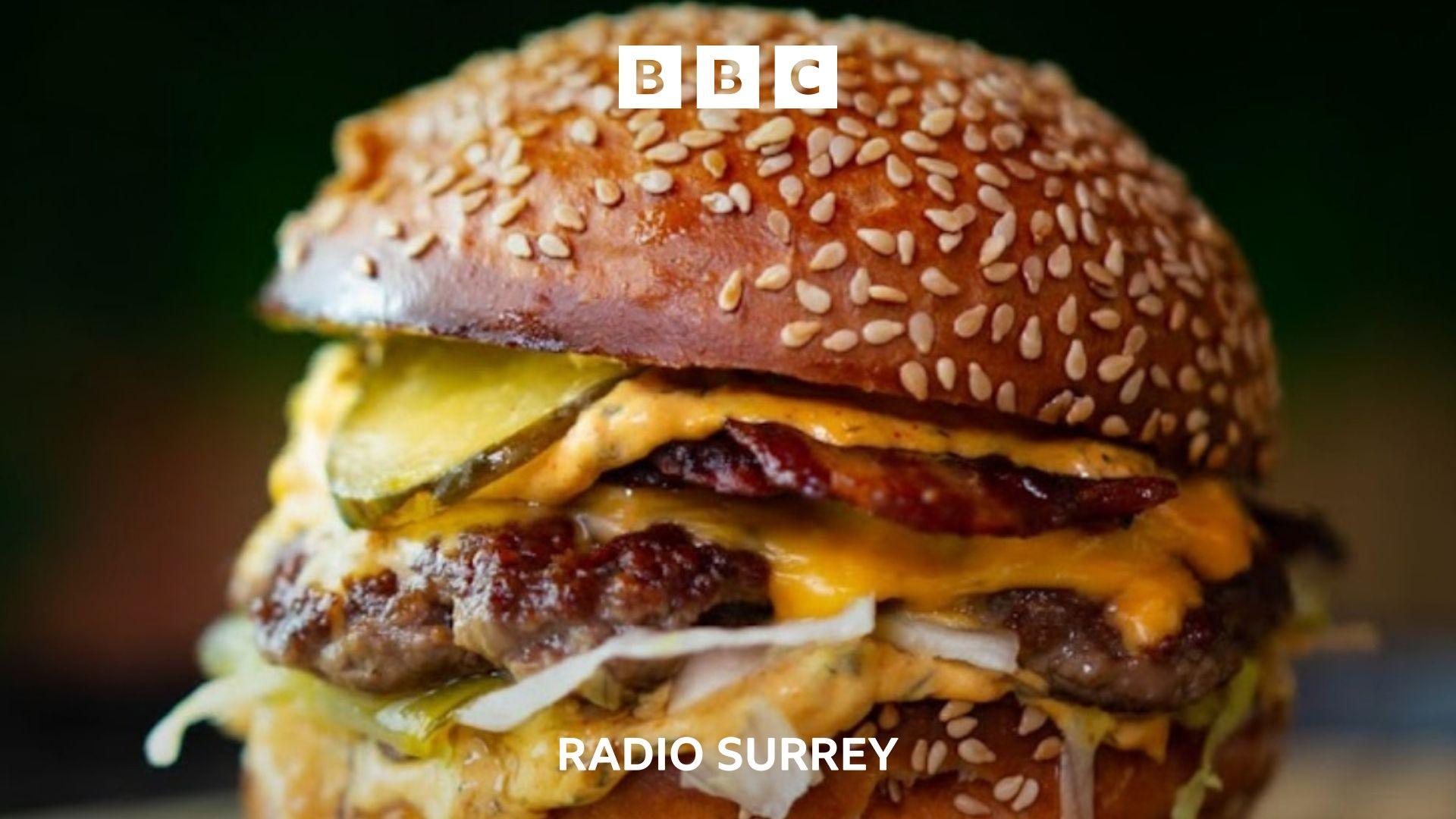 A close up of a juicy beef burger in a sesame seed bun. The burger lies on a bed of lettuce and sauce and is topped with melted cheese, pickles and crispy bacon.