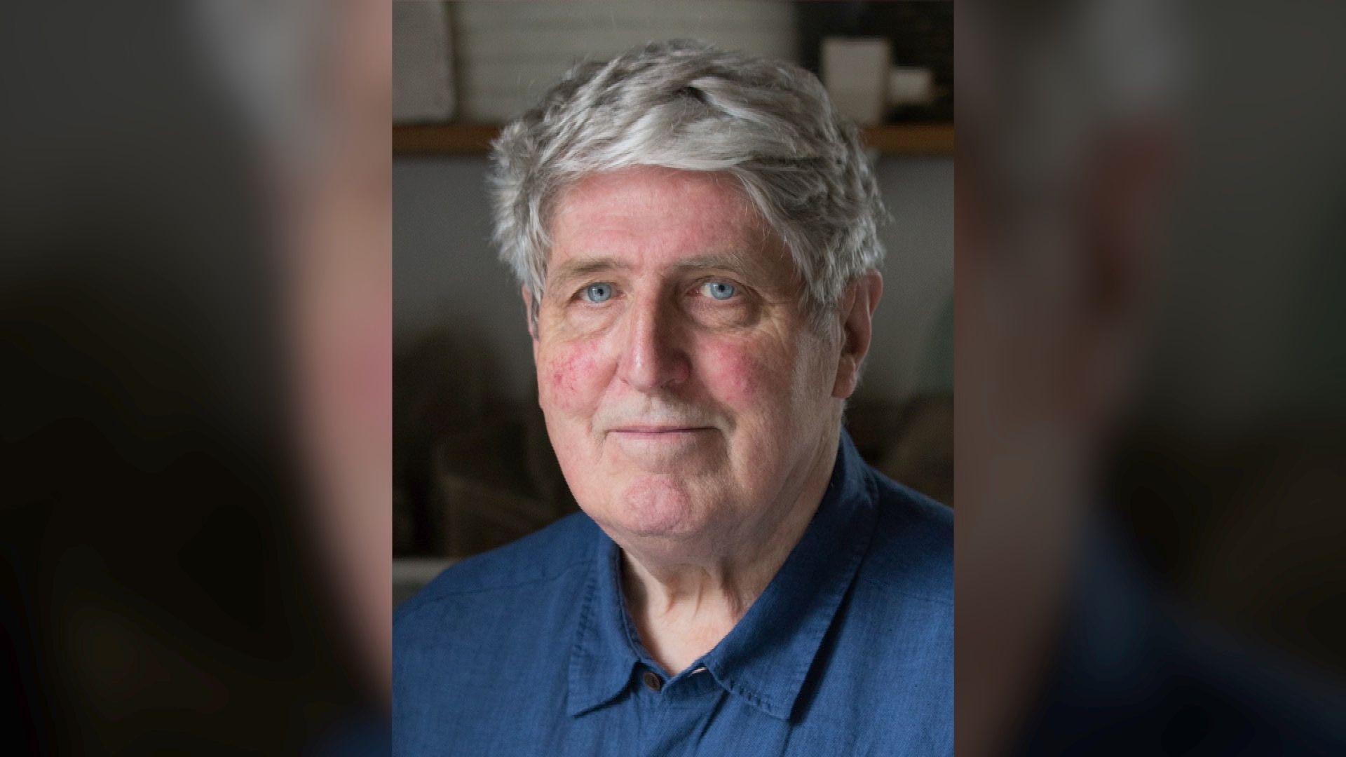 John Maine looks directly at the camera as he poses for a headshot. He has short grey hair and blue eyes. He is wearing a french blue linen collared shirt and the background behind him is blurred.