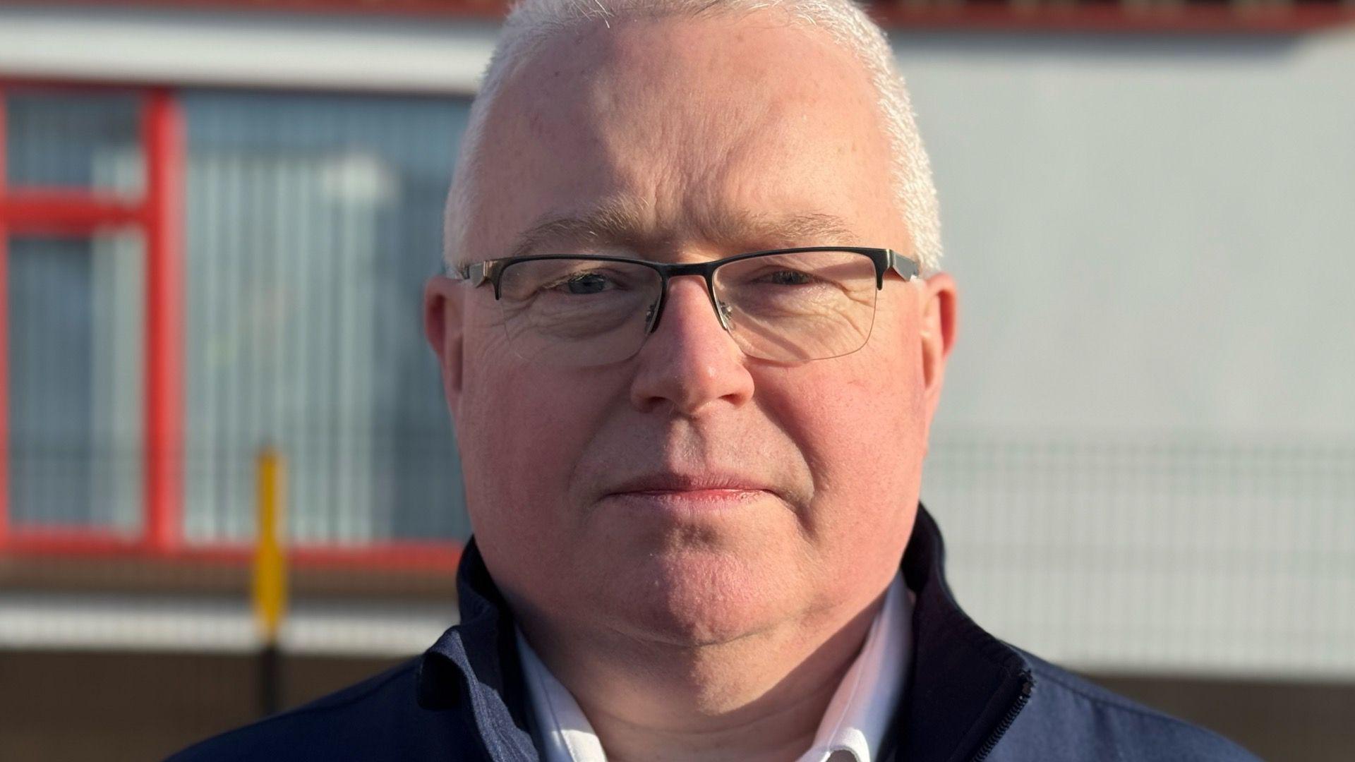 Dermot O'Brien is wearing black glasses. He has a white shirt and navy jacket on. He has short grey hair. The background is blurry.