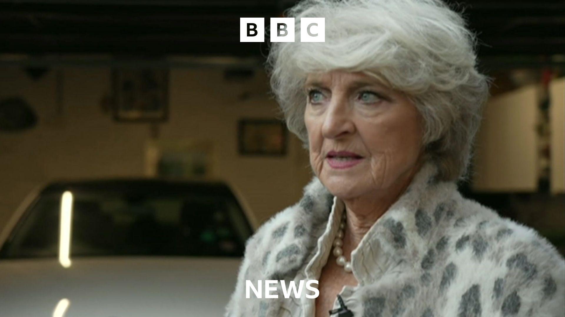 A woman stands in front of her garage. She is well dressed, has grey hair and a string of pearls.