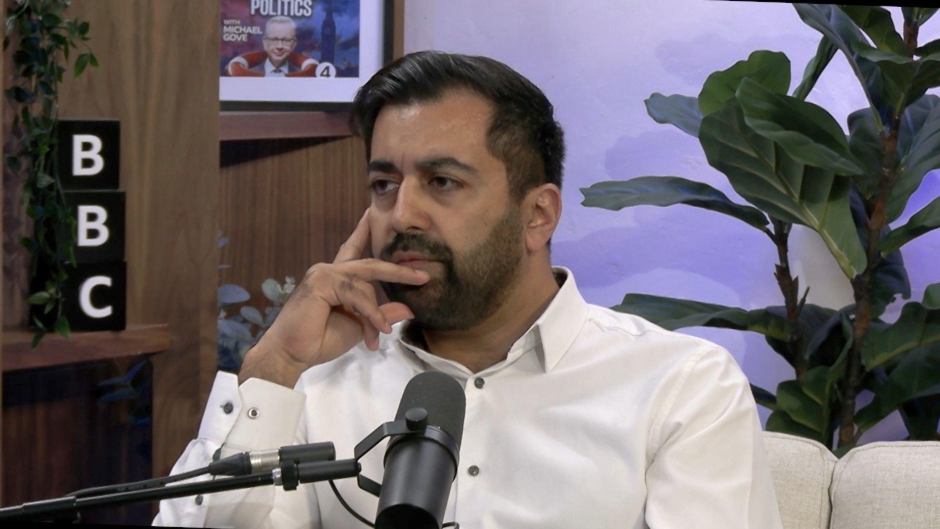 Humza Yousaf in an open-necked white shirt sits at a microphone on a BBC radio studio set. He has his hands against the side of his face as he listens to the question