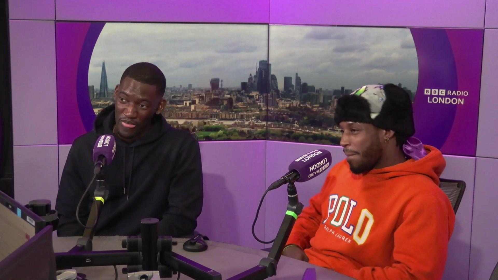 Malachi Kirby (left) in a black hoodie, and Francis Lovehall (right) in a red hoodie and black and grey hat, in the BBC radio London studio behind purple mics as they are interviews. In the background, purple panels and a panoramic image of the London skyline can be seen