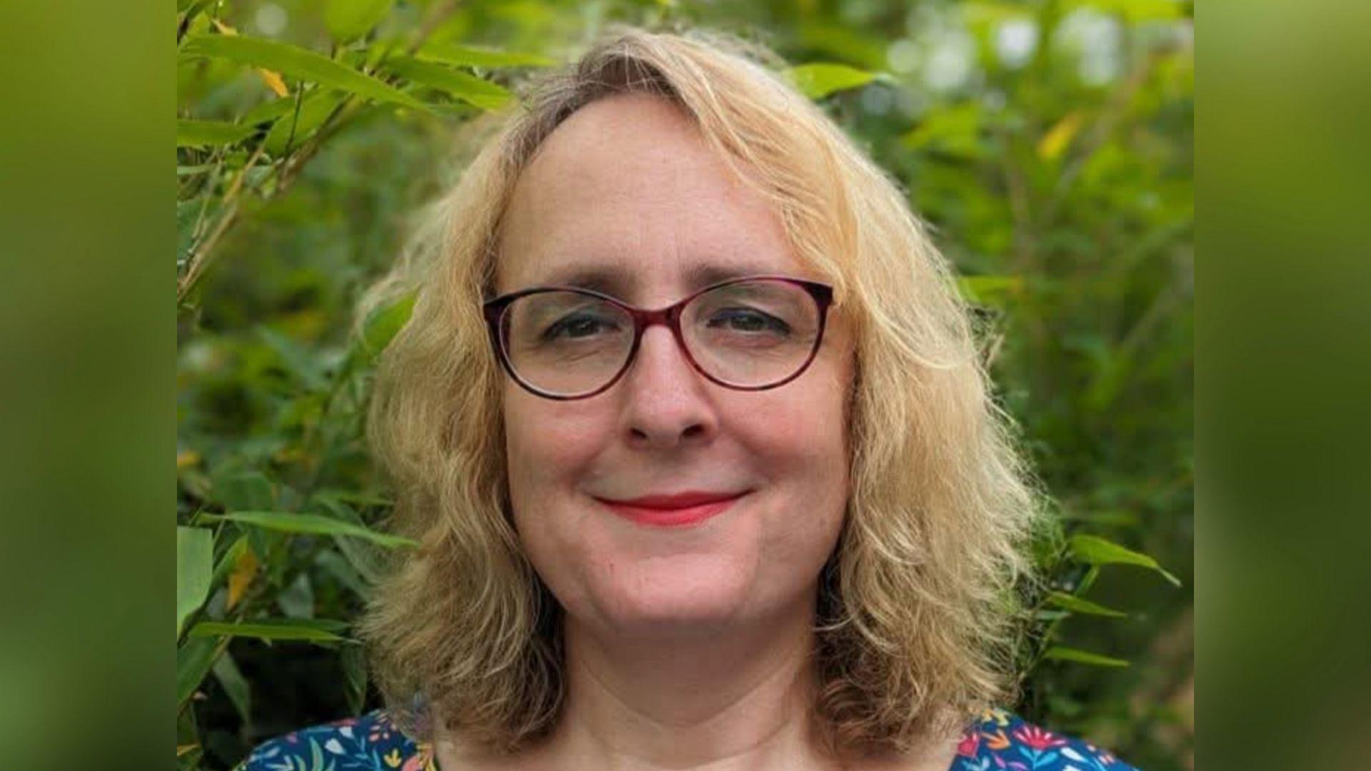 Abby Smith, a woman wearing glasses and a patterned top, is standing against foliage. She is smiling for the camera.