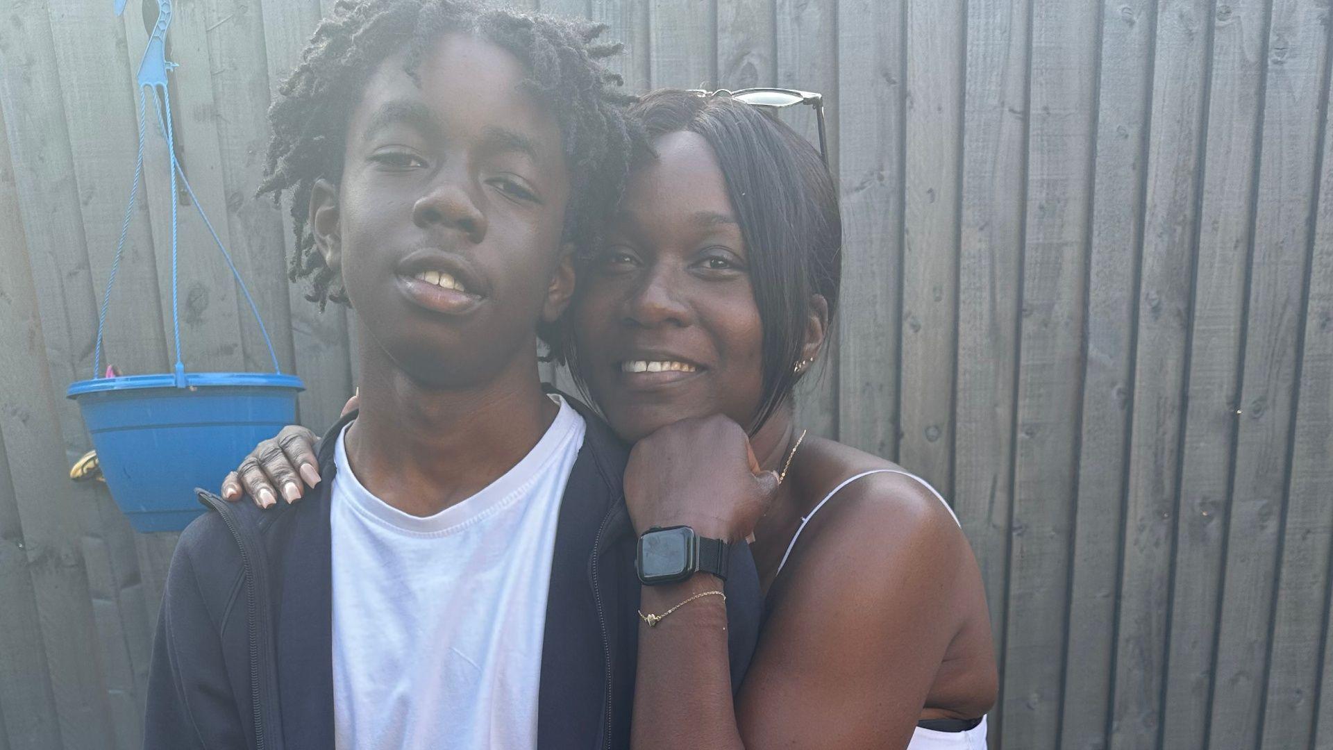 A mother and son pose together in a garden in front of a wooden fence, with the mother standing behind her son and resting an arm on his shoulder.