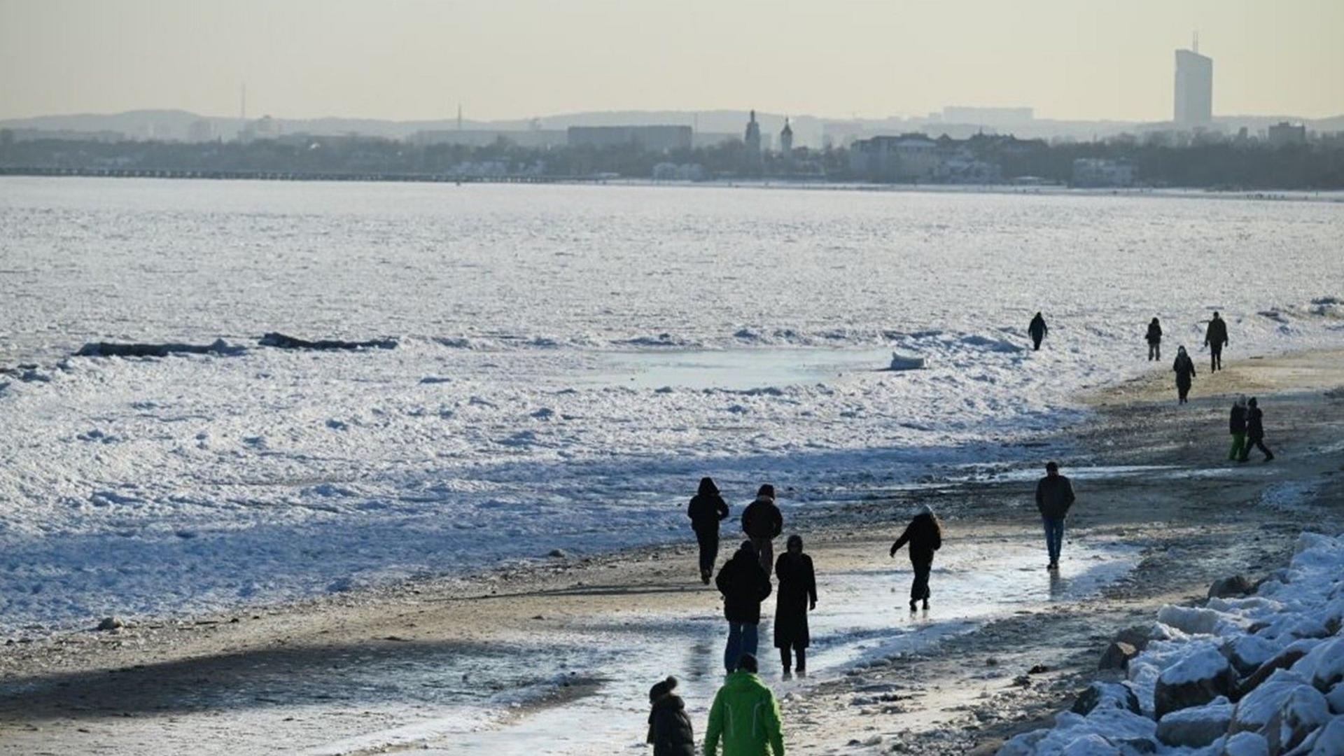 People walking along the beach in cold weather with ice covered seas.