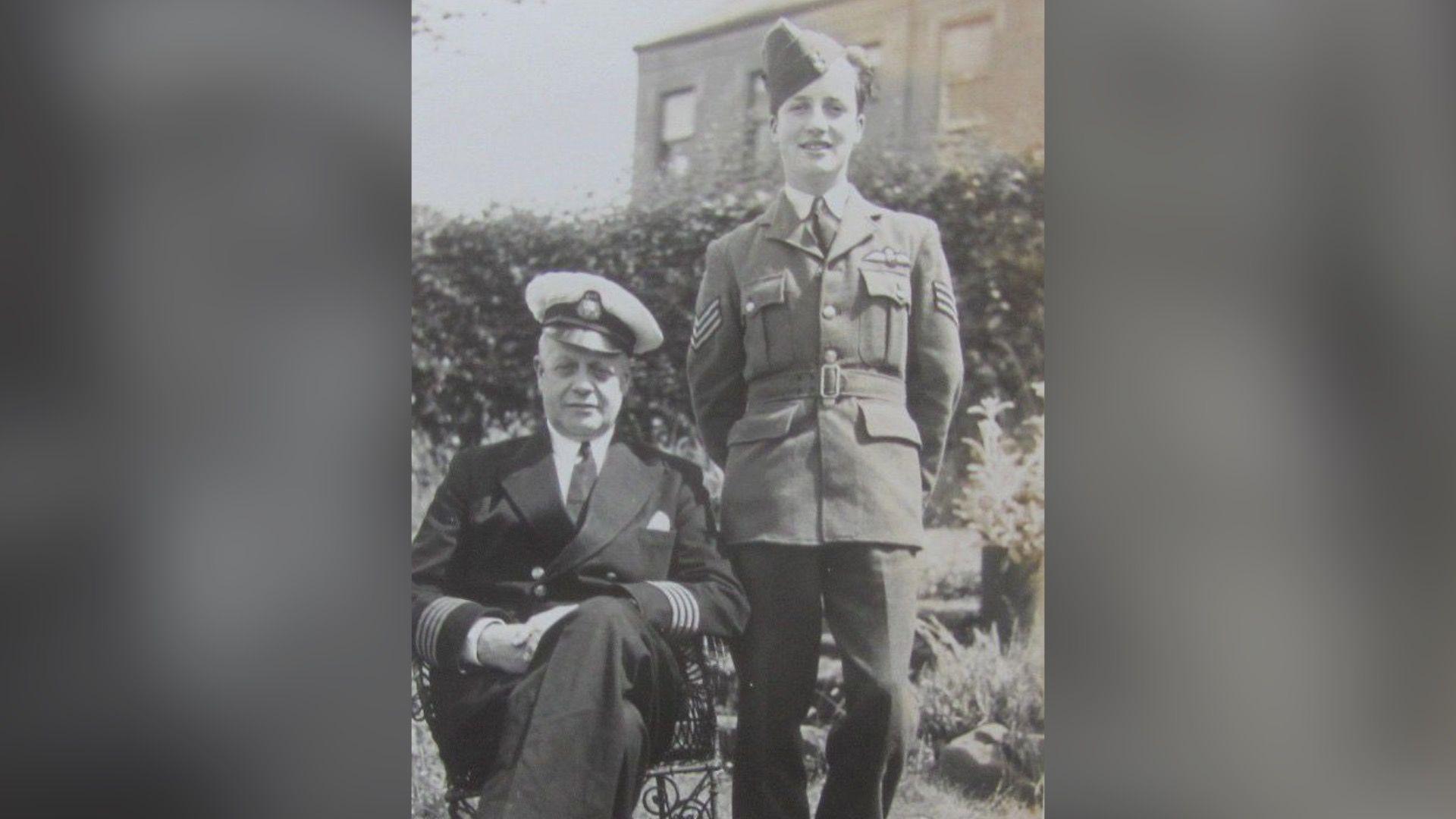 A young man in his military uniform stands beside an old man in uniform who is sitting down. They are in a garden.