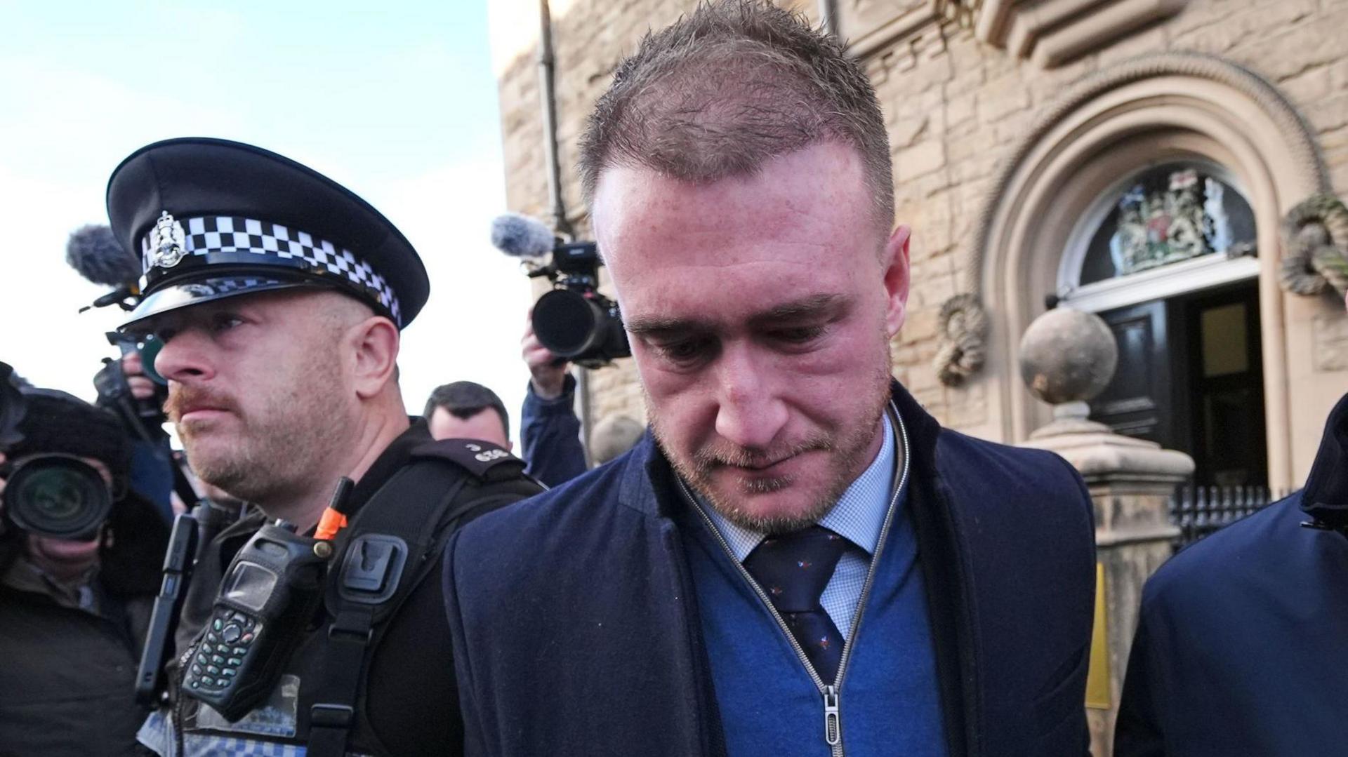 Rugby star Stuart Hogg with his head bowed in a zip top with a shirt and tie underneath stands outside Selkirk Sheriff Court flanked by a police officer