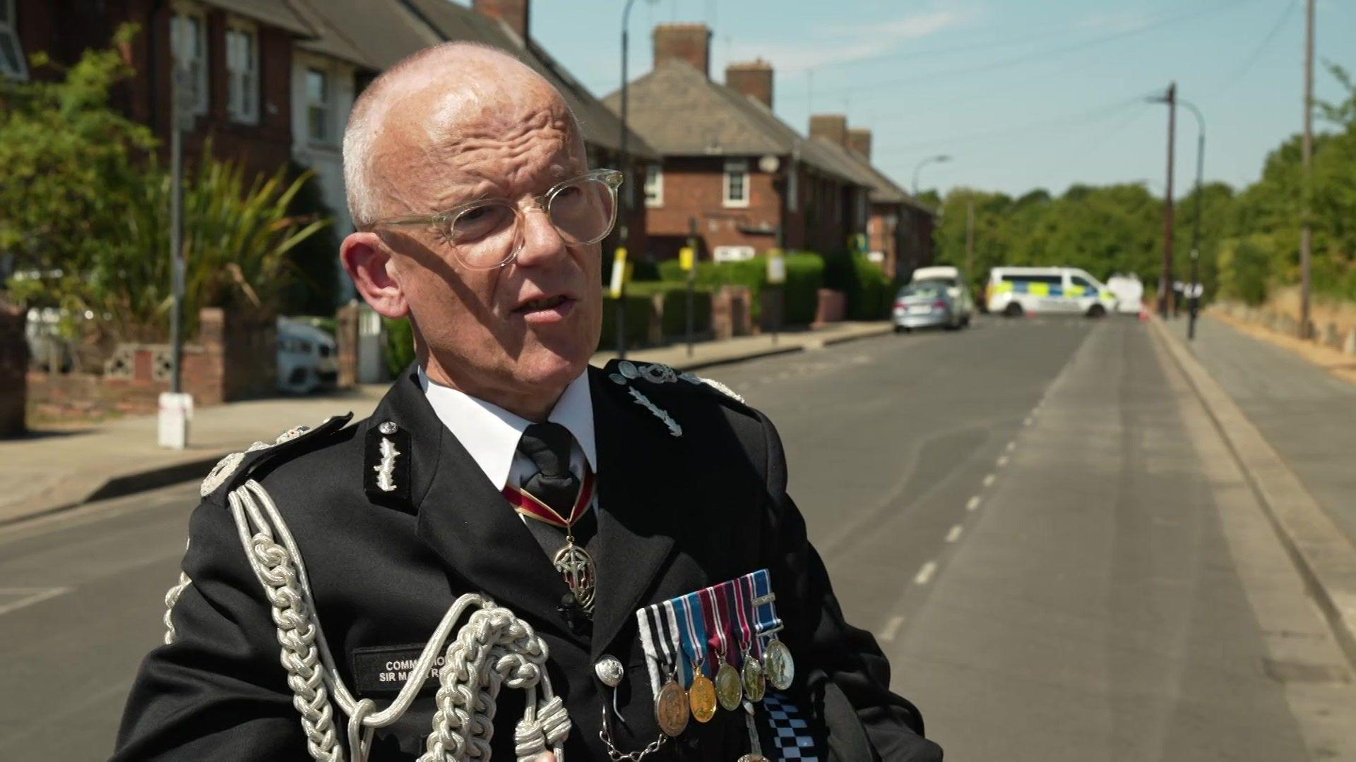 Metropolitan Police Commissioner Sir Mark Rowley speaking outdoors on a residential street. He is wearing full dress uniform complete with silver aiguillettes, medals, and clear-framed glasses, with police vehicles and brick terraced houses visible in the blurred background.