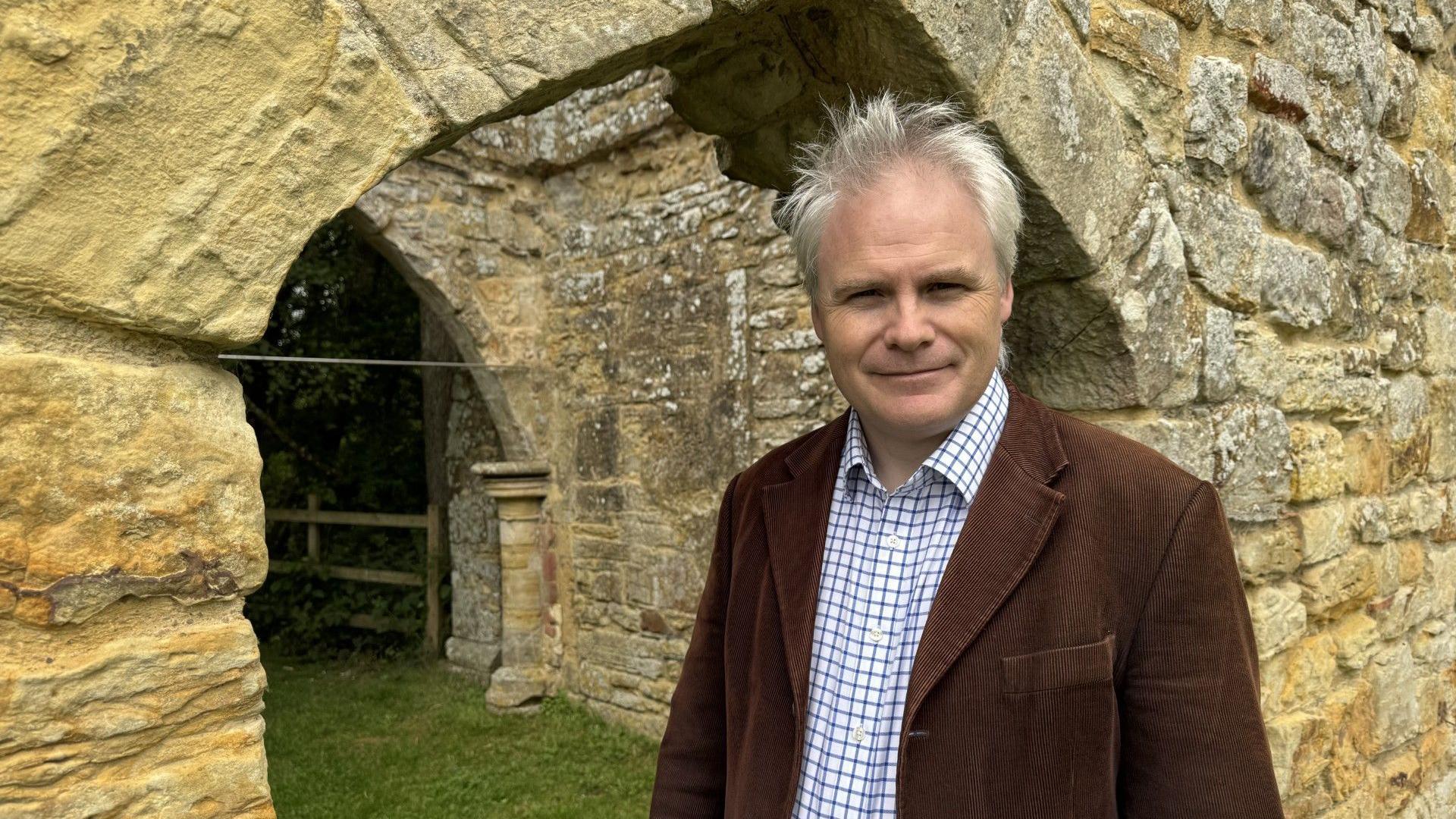 Roy Porter stands in front of the abbey ruins which are in a pale stone. He is looking at the camera and wearing a blue and white checked shirt with a brown jacket.