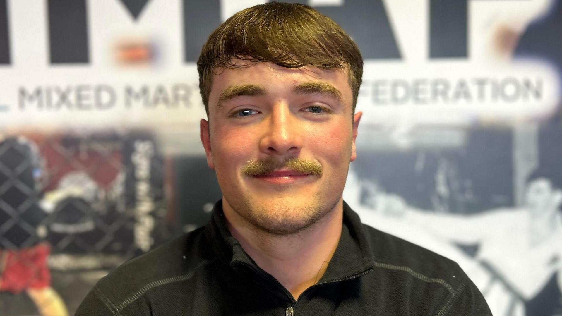 Ruairi O'Kane has brown hair and a moustache. He is smiling at the camera, wearing a black t-shirt.