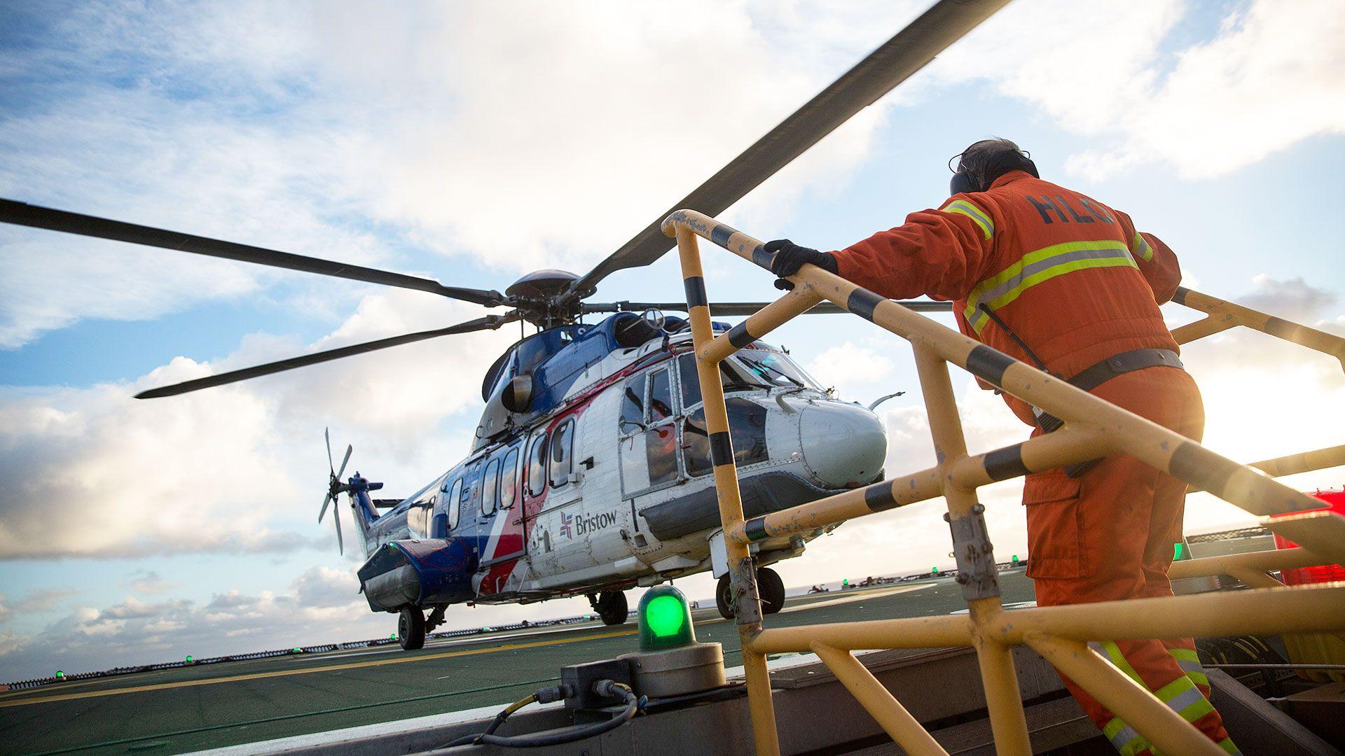A helicopter lands on an oil rig and a man in orange overalls is climbing up steps and heading towards it
